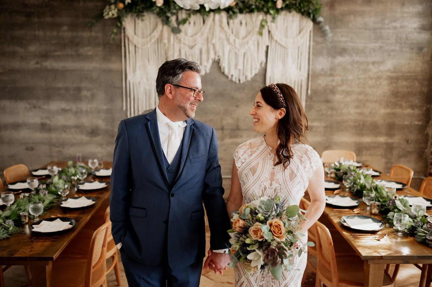 A couple in wedding attire smiles at each other while holding hands, with a beautifully decorated table setting at their intimate wedding at Opal 28 in the background.