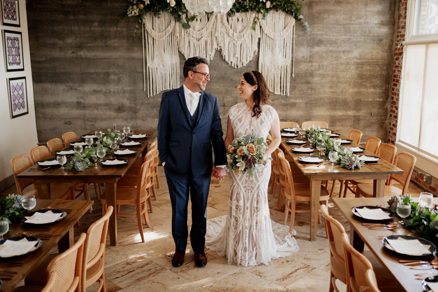 A couple in formal attire stands smiling at each other in a decorated dining area with a bohemian backdrop, celebrating their wedding at Opal 28.