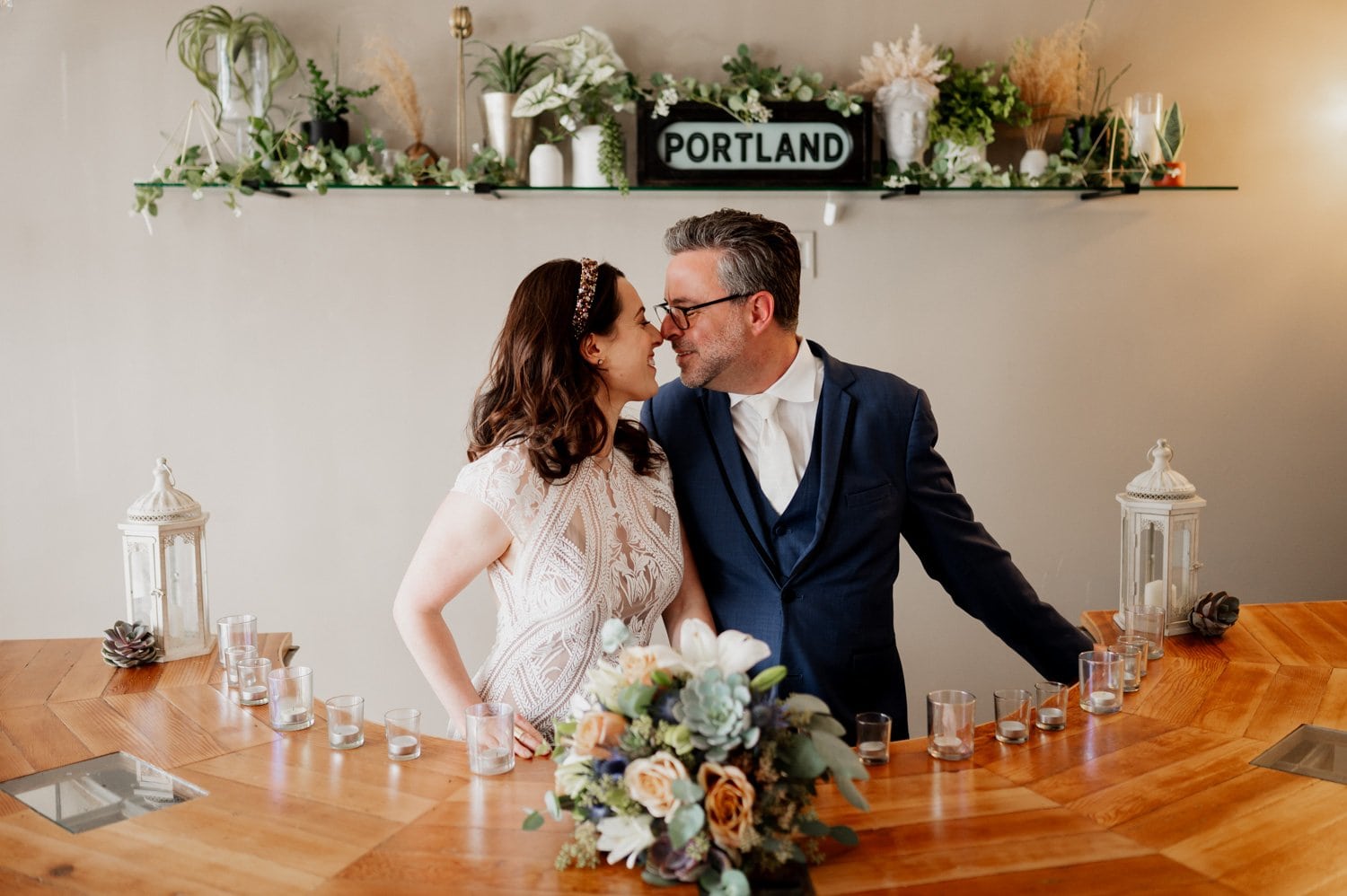 A couple in wedding attire smiles at each other over a bar decorated with candles and flowers during their wedding celebration at Opal 28.