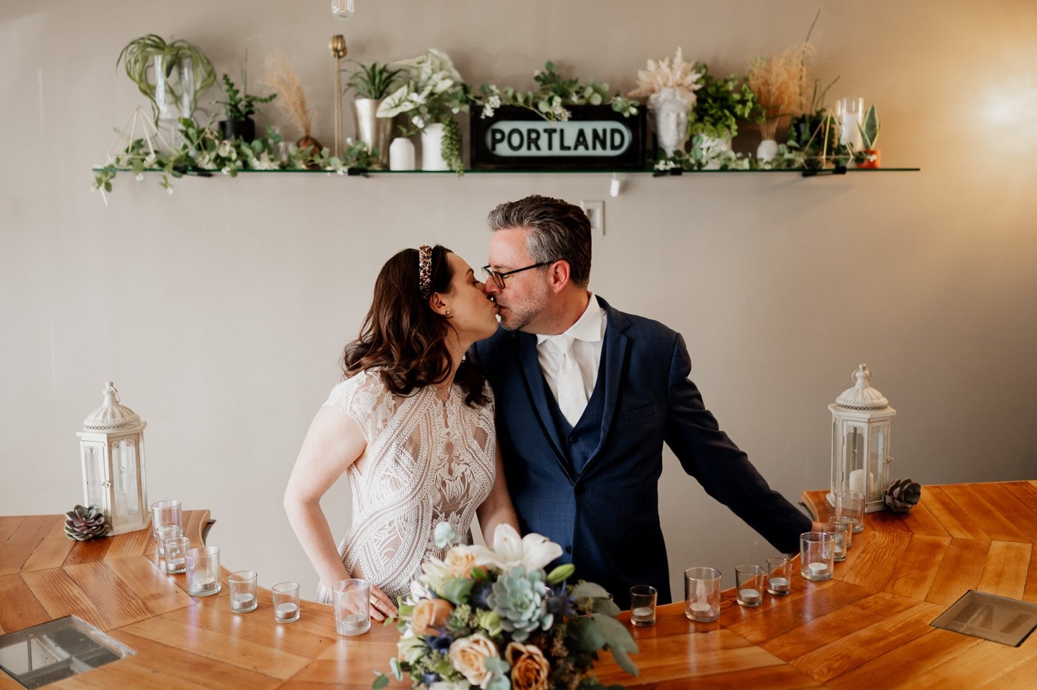 A couple shares a kiss at their wedding at Opal 28, standing behind a wooden table decorated with candles and flowers, under a glowing PORTLAND sign.
