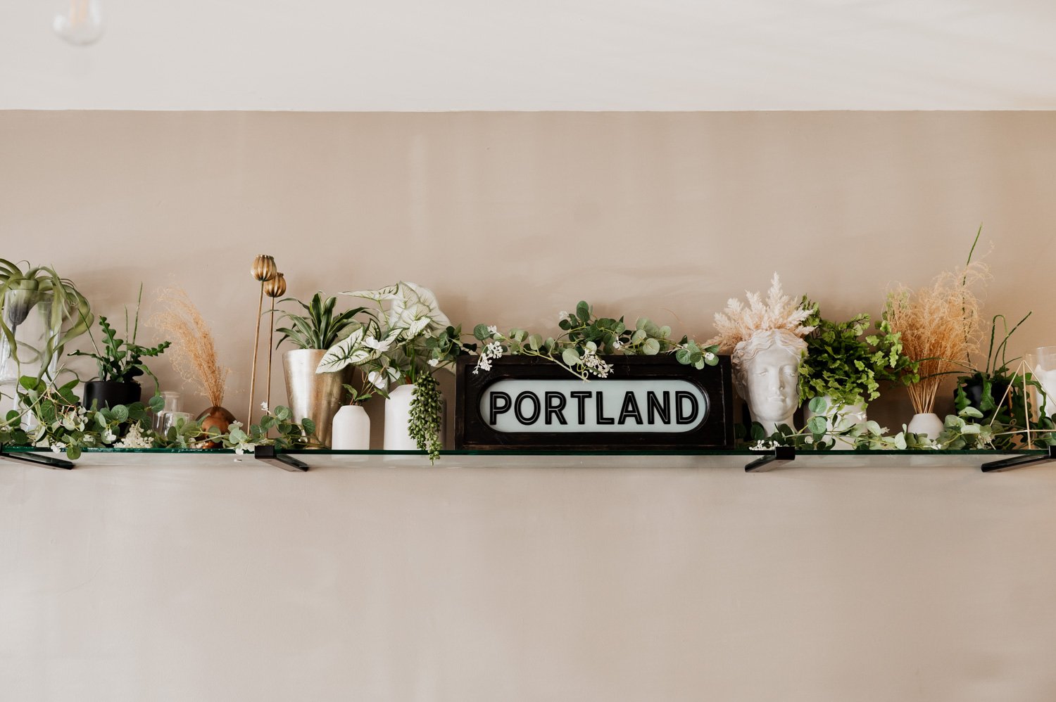 A shelf decorated with lush plants and elegant vases, next to a Portland sign, set against a soothing beige wall.