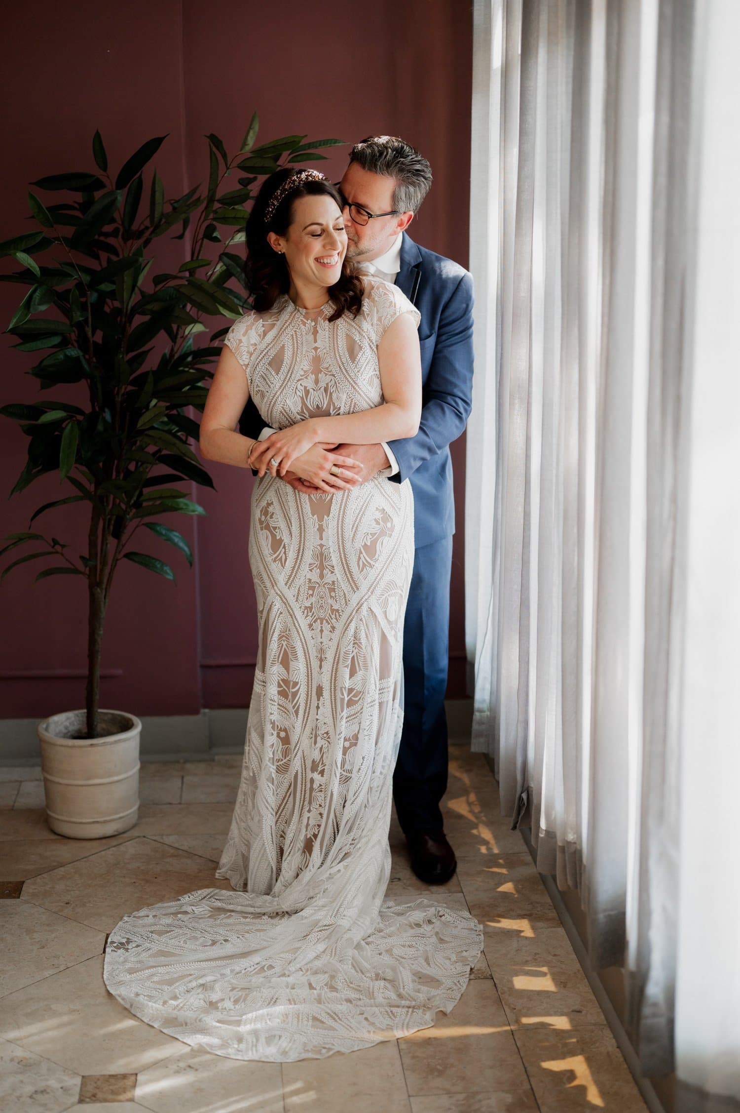 A couple embraces by a window at Opal 28; the woman wears a white lace gown and the man is dressed in a blue suit.