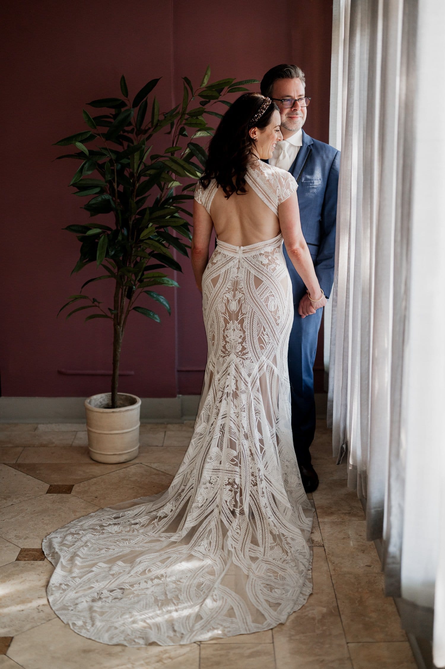 A bride in a detailed lace gown and a groom in a blue suit stand by a window, holding hands, with a potted plant close to them at their wedding at Opal 28.