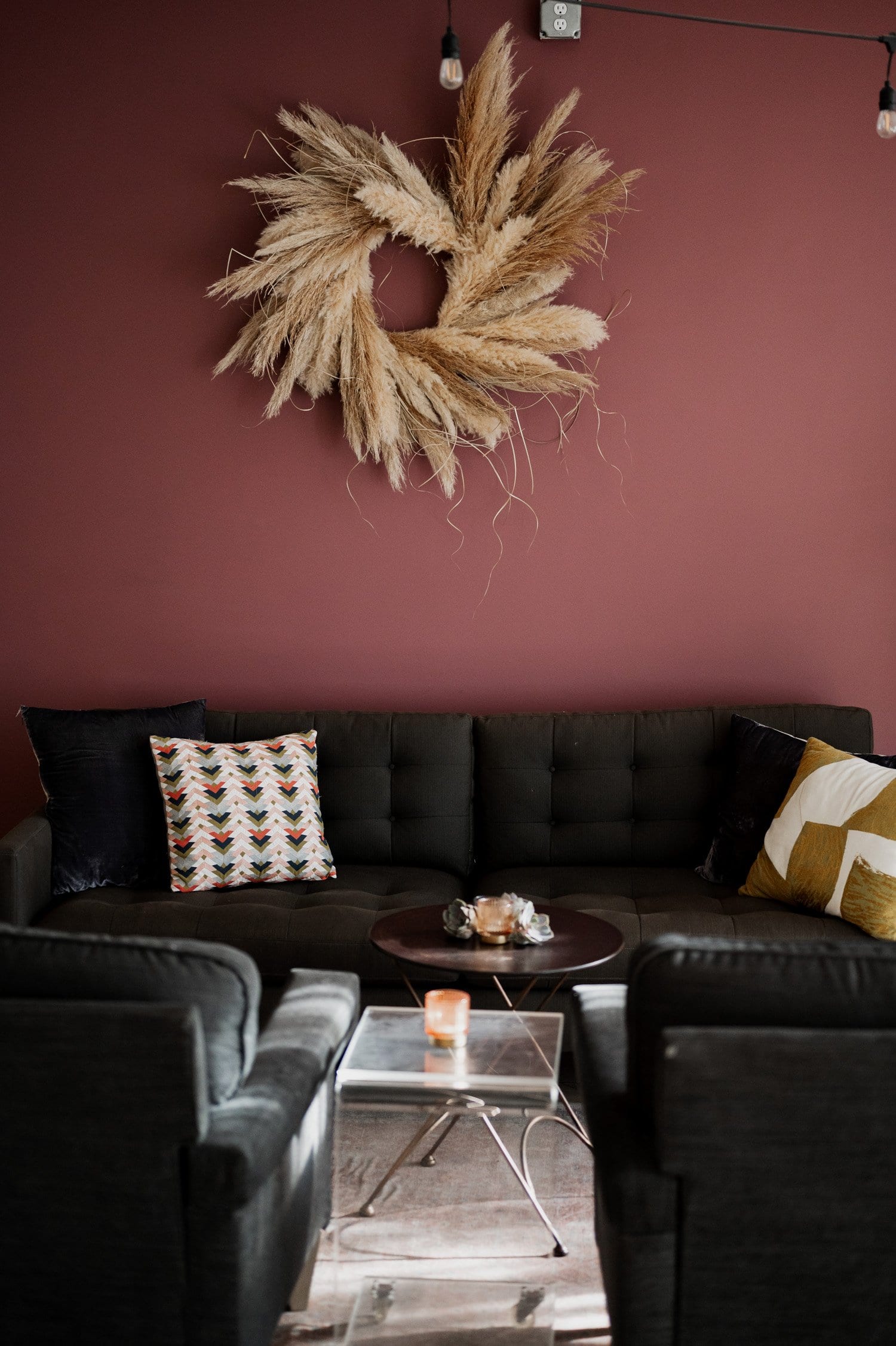 A modern living room with a dark sofa, abstract-patterned cushions, a pampas grass wreath on the maroon wall, and a chic coffee table.