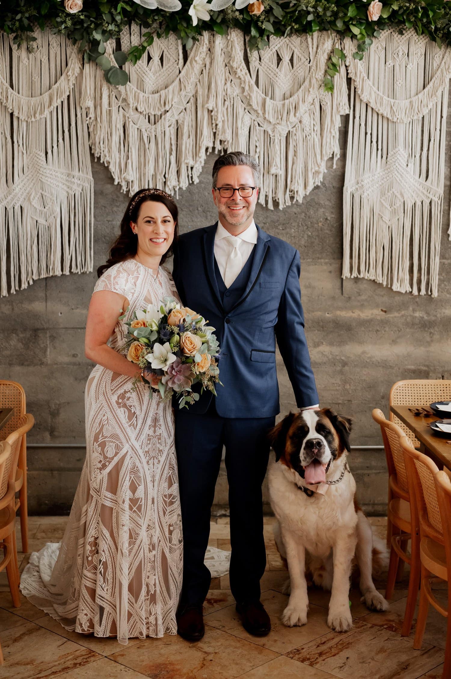 A couple wearing wedding attire poses with their large dog in front of a macramé backdrop at Opal 28.