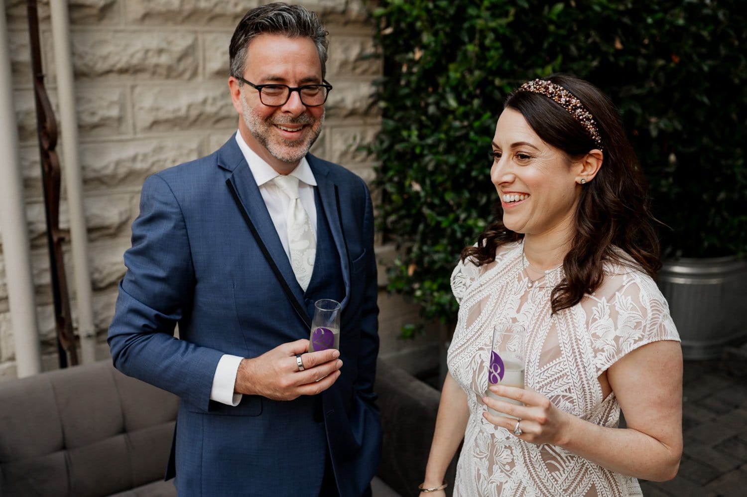 A well-dressed couple smiles brightly while holding drinks outdoors by a stone wall and lush greenery, capturing a joyful moment at Opal 28.