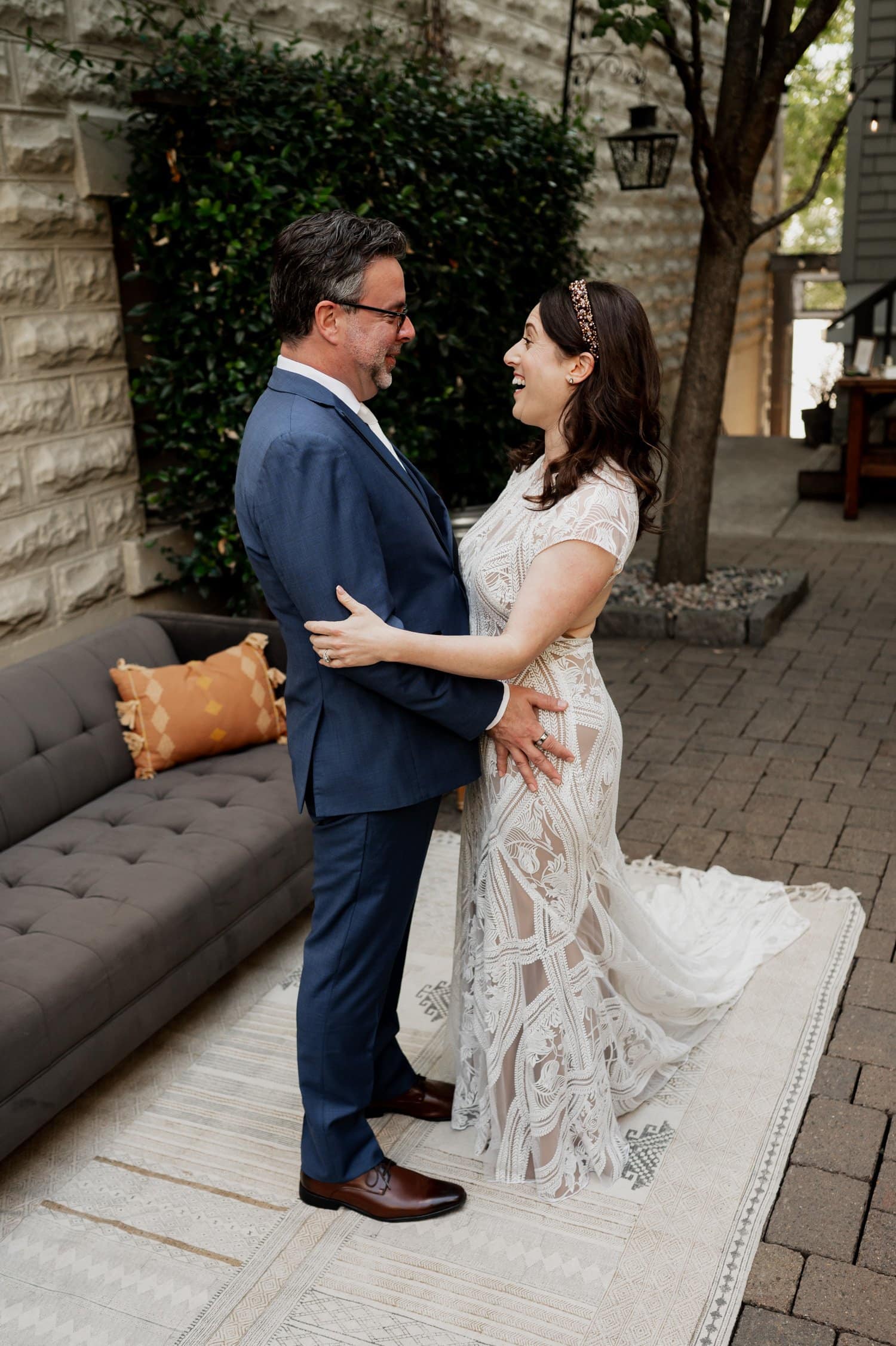 A couple in formal attire smiles at each other during their wedding at Opal 28, with a sofa and lush greenery in the charming outdoor setting.