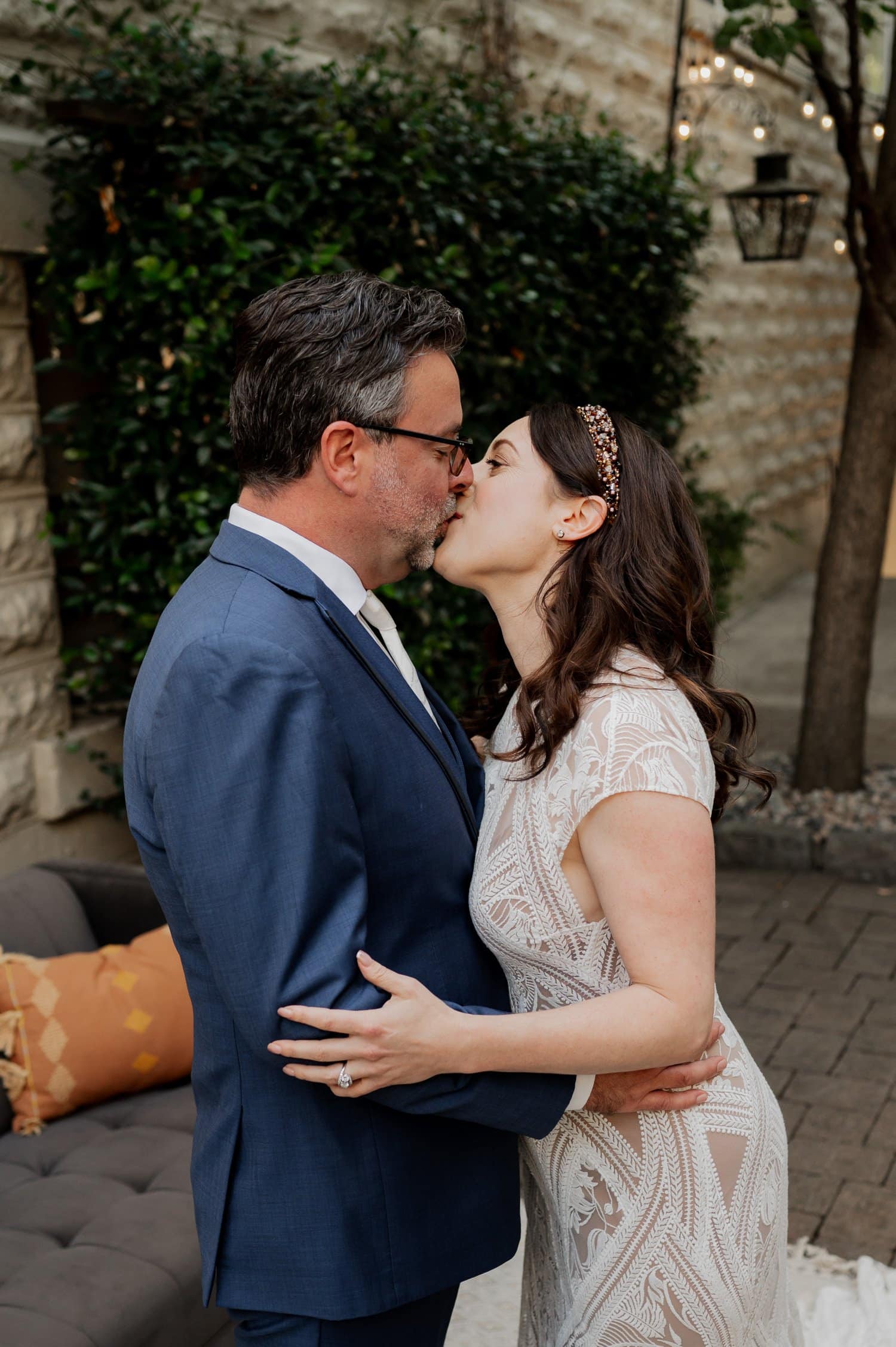 The couple, dressed in elegant formal attire, shares a romantic kiss outdoors near a brick wall with lush green foliage during their wedding at Opal 28.