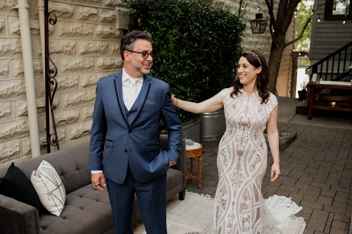 A bride in a white gown touches the grooms shoulder as he stands beside her in a blue suit, with a stone wall and lush greenery surrounding them at their wedding at Opal 28.