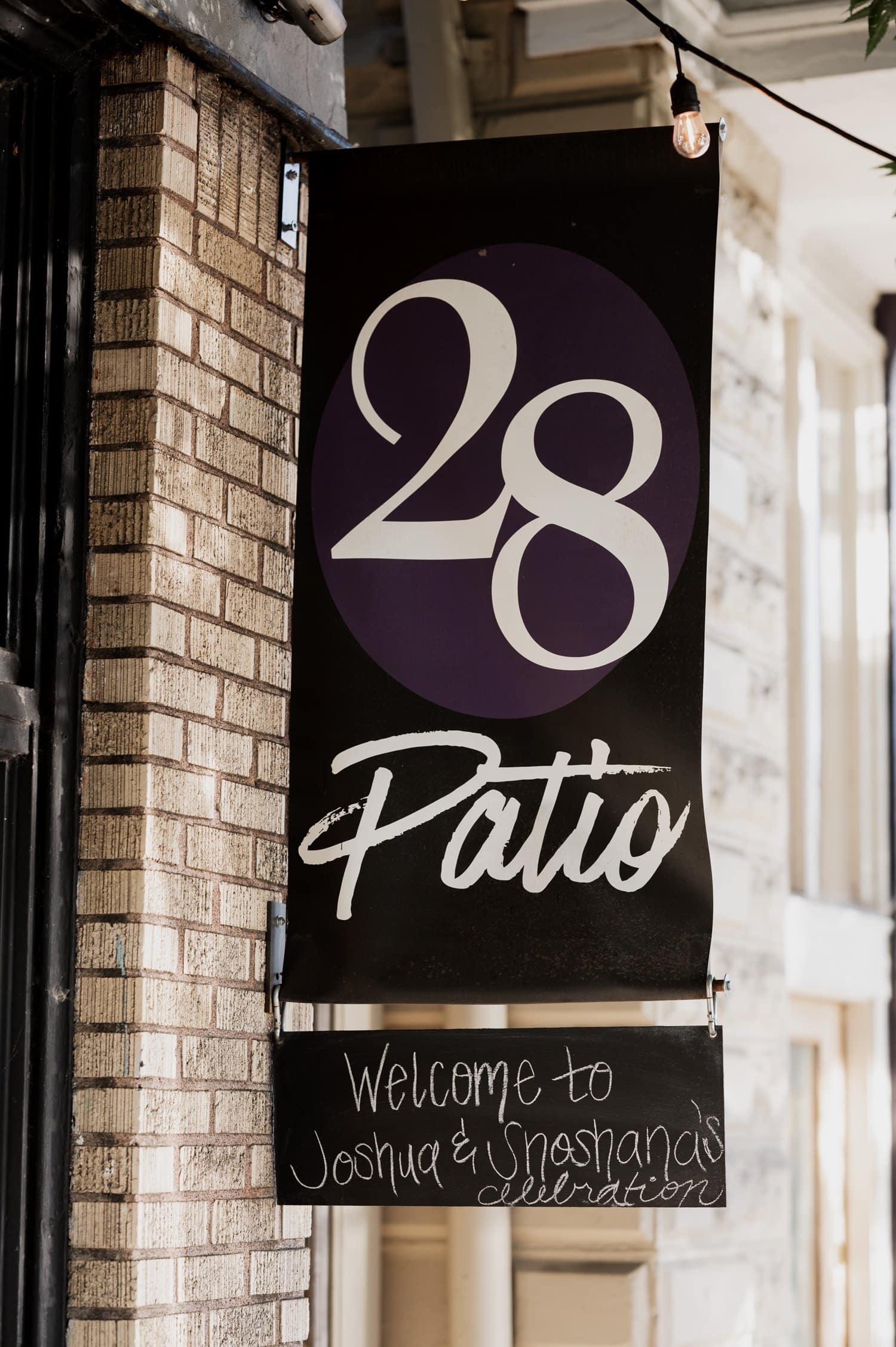 A sign reading 28 Patio with the text Welcome to Joshua & Uneshanas celebration elegantly hangs on a brick wall, marking their wedding at Opal 28.