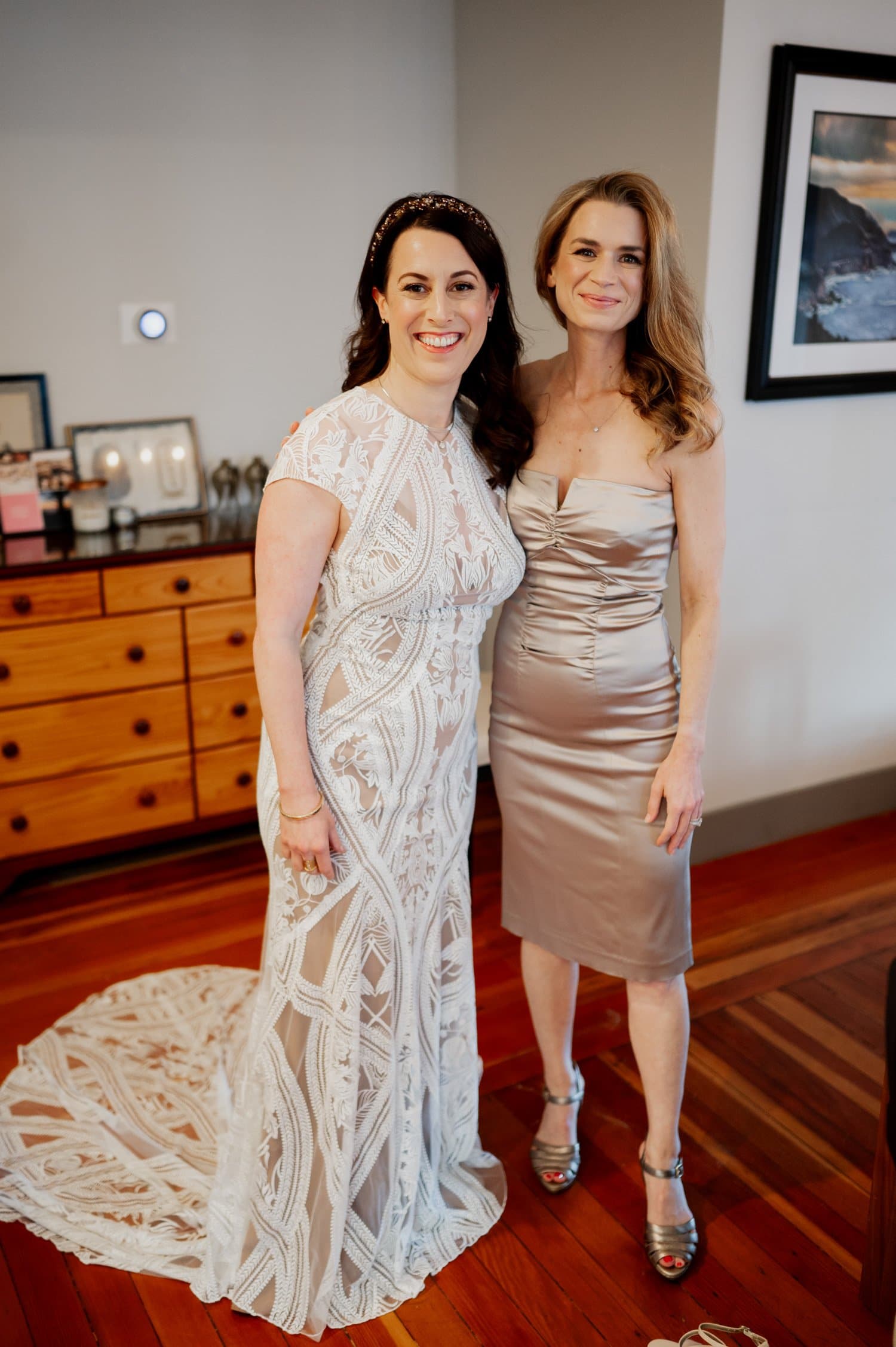Two women smiling; one in a white lace wedding dress and the other in a silver dress, standing together indoors. Theyre getting ready at a home in NE Portland before heading to Opal 28 for the wedding.