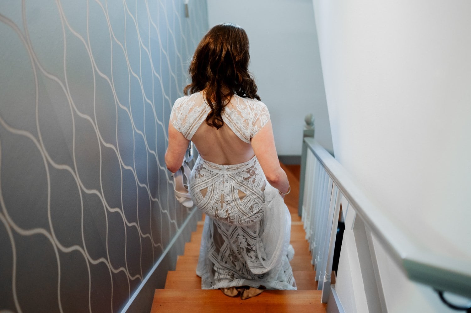 A woman in an elegant white dress gracefully descends a staircase against a wavy patterned wall, getting ready at a home in NE Portland before her wedding at Opal 28.