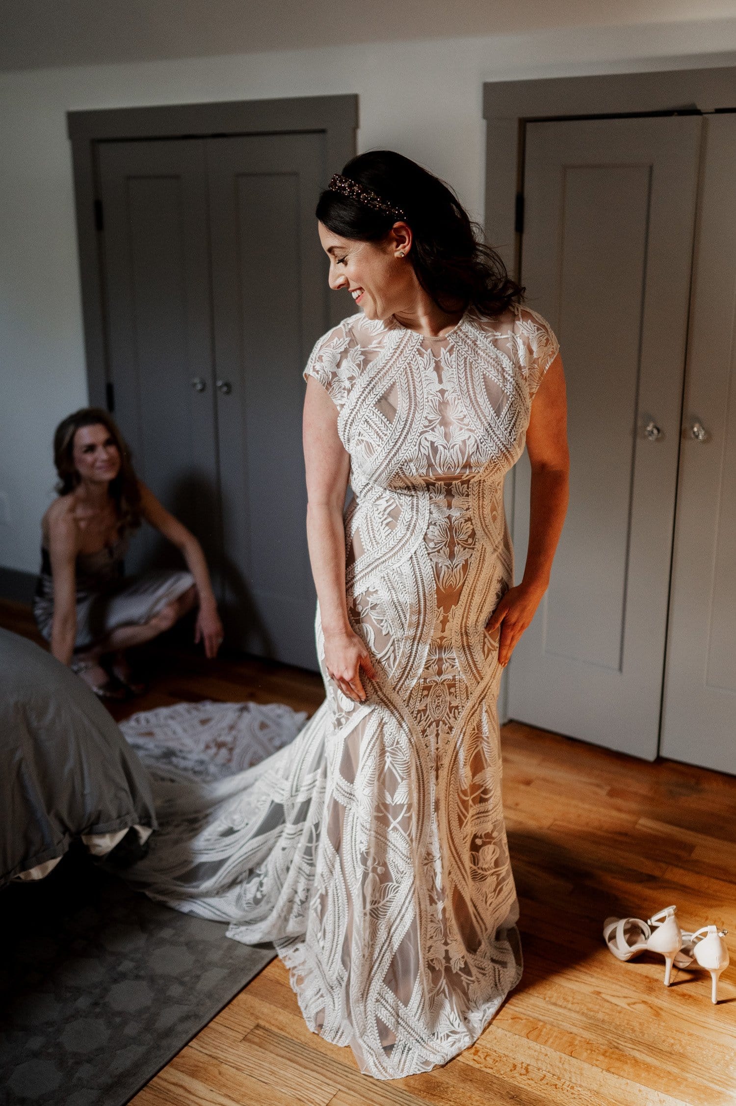 In a charming NE Portland bedroom, the bride in an ornate gown smiles at a seated woman, shoes scattered on the floor nearby, as they savor the precious moments of getting ready before heading to their wedding celebration at Opal 28.