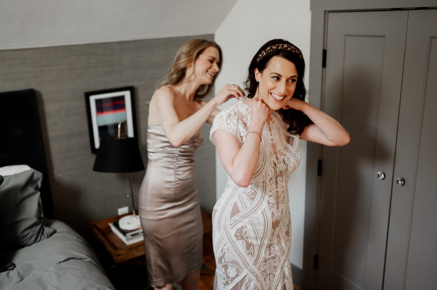 In a cozy NE Portland bedroom, two women share smiles as they prepare for the big day at Opal 28. One gently adjusts the others lace dress near a bed and nightstand, capturing a heartfelt moment of getting ready before the wedding.