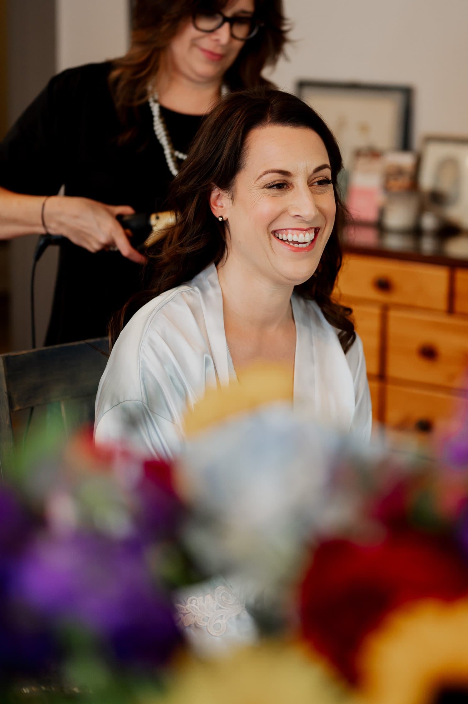 A woman smiles as her hair is styled amidst colorful flowers in the foreground, getting ready at a home in NE Portland before a wedding at Opal 28.