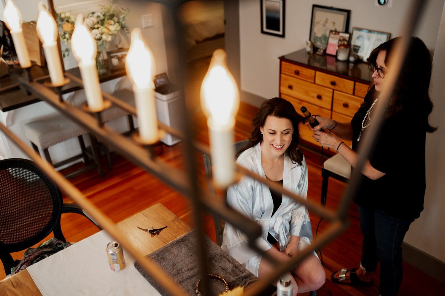 A woman getting her hair styled, sitting in a cozy room with soft lighting and a table with drinks and accessories nearby, is getting ready at a home in NE Portland before a wedding at Opal 28.