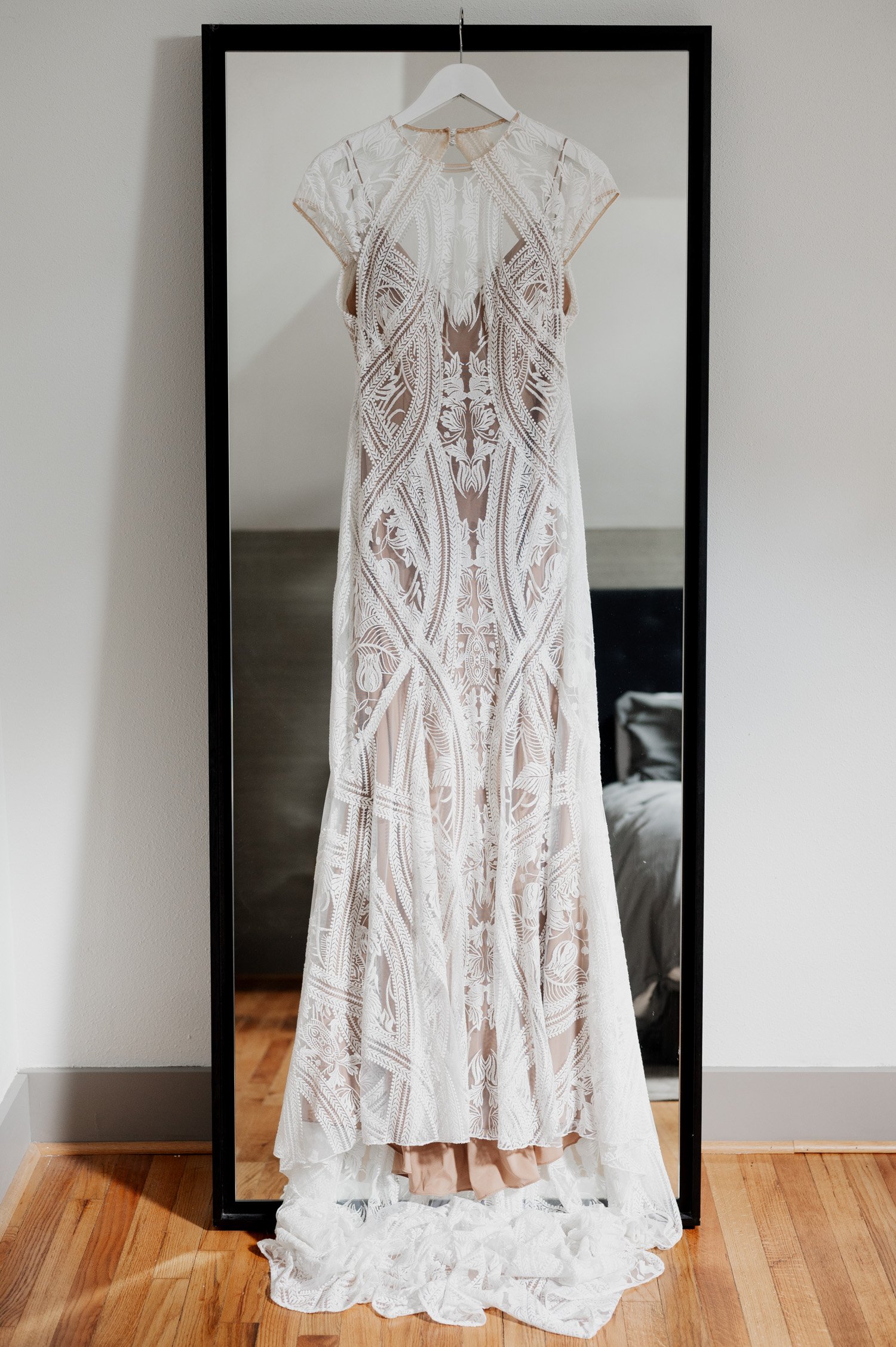 An ivory lace wedding dress with intricate patterns hangs gracefully on a mirror, reflecting the excitement of getting ready at a home in NE Portland before heading to Opal 28 for the big day. The soft wood floor beneath adds warmth to this moment of anticipation.
