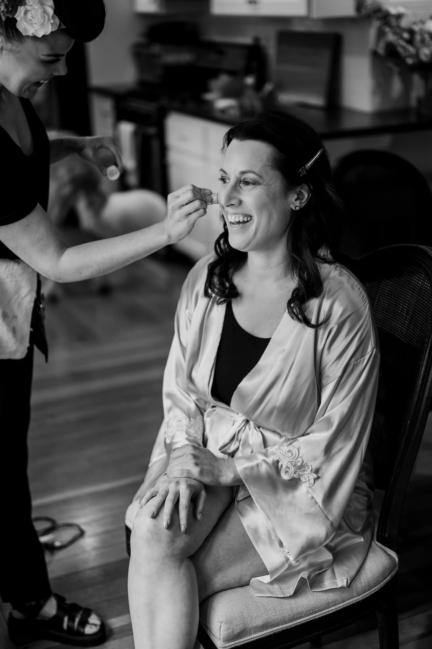 A bride smiles as she gets her makeup done, sitting in a kitchen chair wearing a silk robe. This black and white photo captures her getting ready at a home in NE Portland before her wedding at Opal 28.