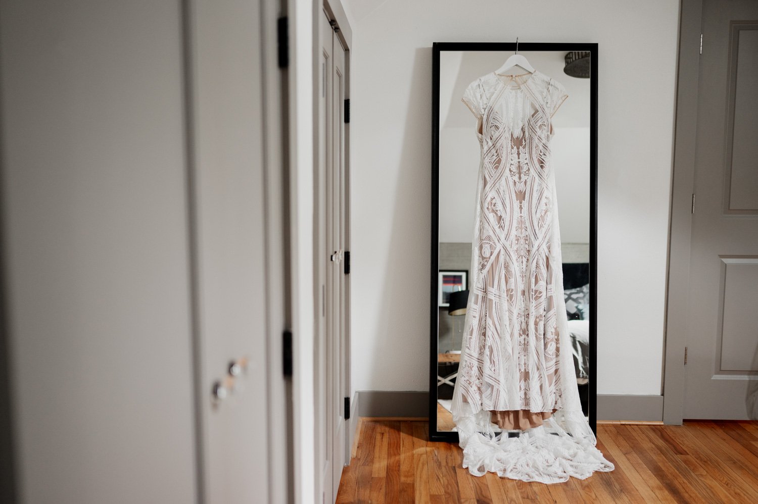 A long, lacy white dress hangs on a mirror in a cozy room with wooden floors and gray walls at home in NE Portland before a wedding at Opal 28.