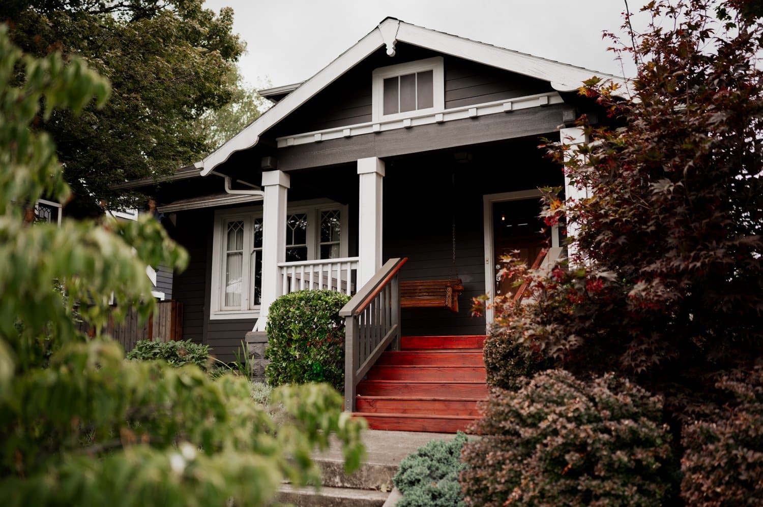 A dark gray house with red steps, nestled among trees and shrubs under a cloudy sky, serves as the perfect backdrop for getting ready at a home in NE Portland before a wedding at Opal 28.