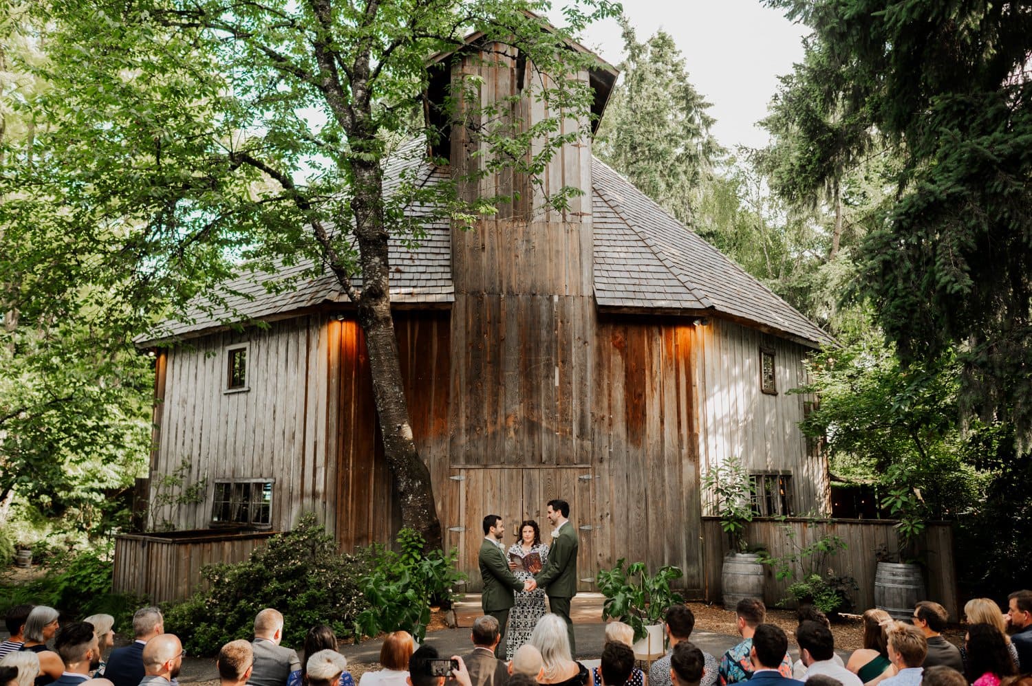 A couple stands before an officiant for their Cornelius Pass Roadhouse wedding beside a rustic wooden, octogonal barn, surrounded by lush greenery. Guests are seated, witnessing the event beneath a clear, bright sky.