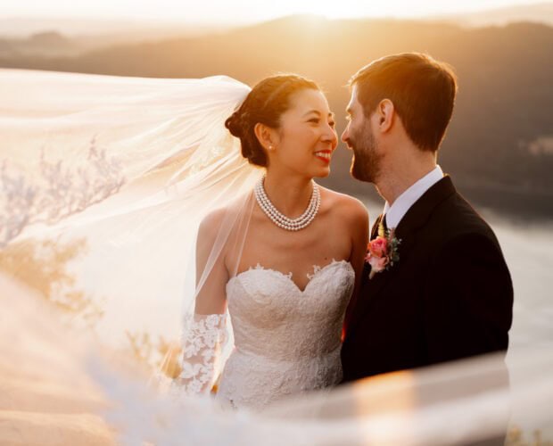 Bride and groom smiling at each other with a flowing veil, overlooking a scenic sunset view at Angels Rest.