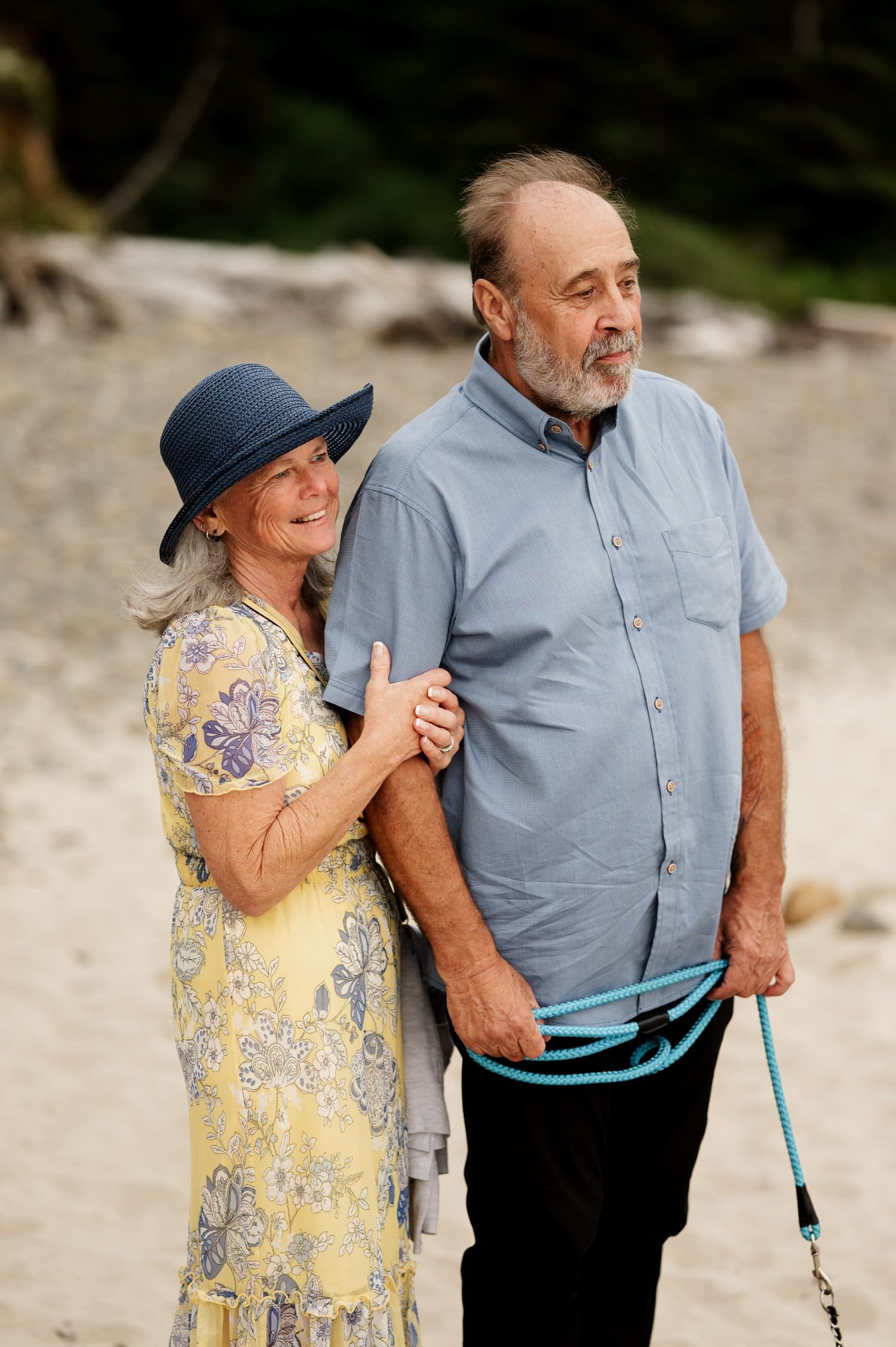 Elderly couple standing on Short Sands Beach, the woman in a floral dress and blue hat smiles, holding the mans arm. Hes in a light blue shirt, clutching a dog leash.
