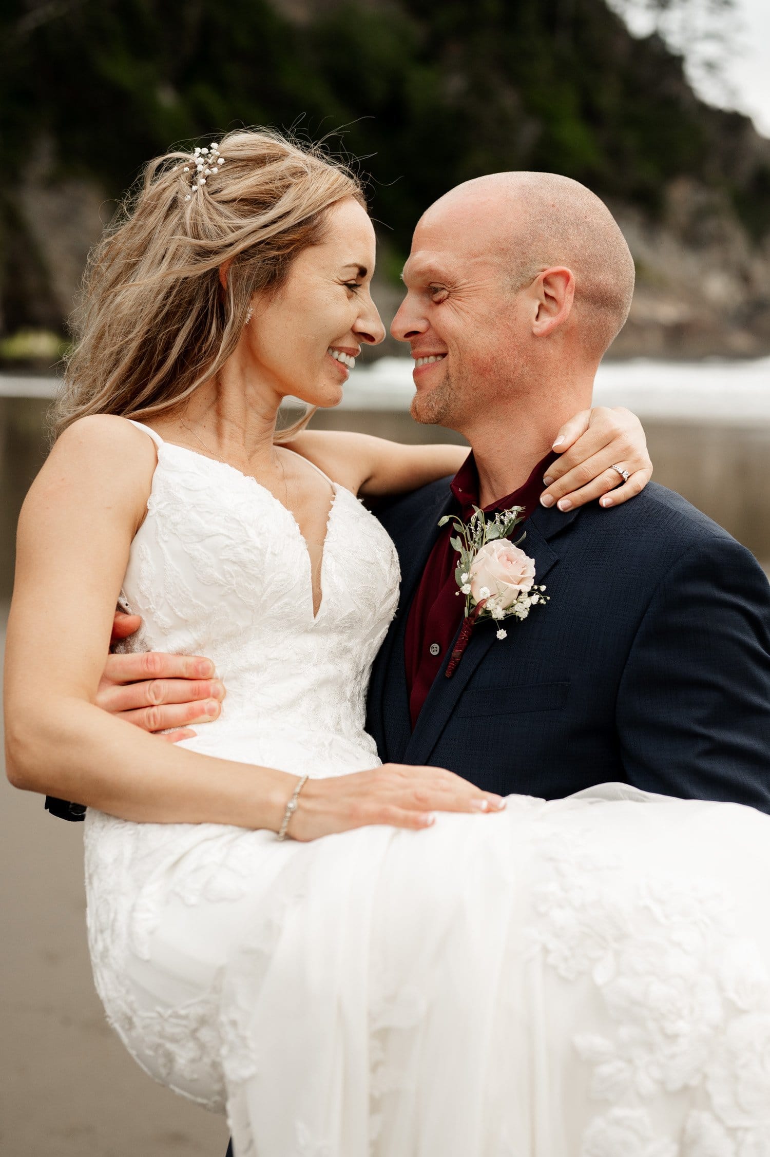A groom in a dark suit joyfully carries his bride, wearing a white wedding dress, at Short Sands Beach. They smile warmly at each other, surrounded by the lush greenery and gentle ocean waves, capturing the essence of an intimate elopement.