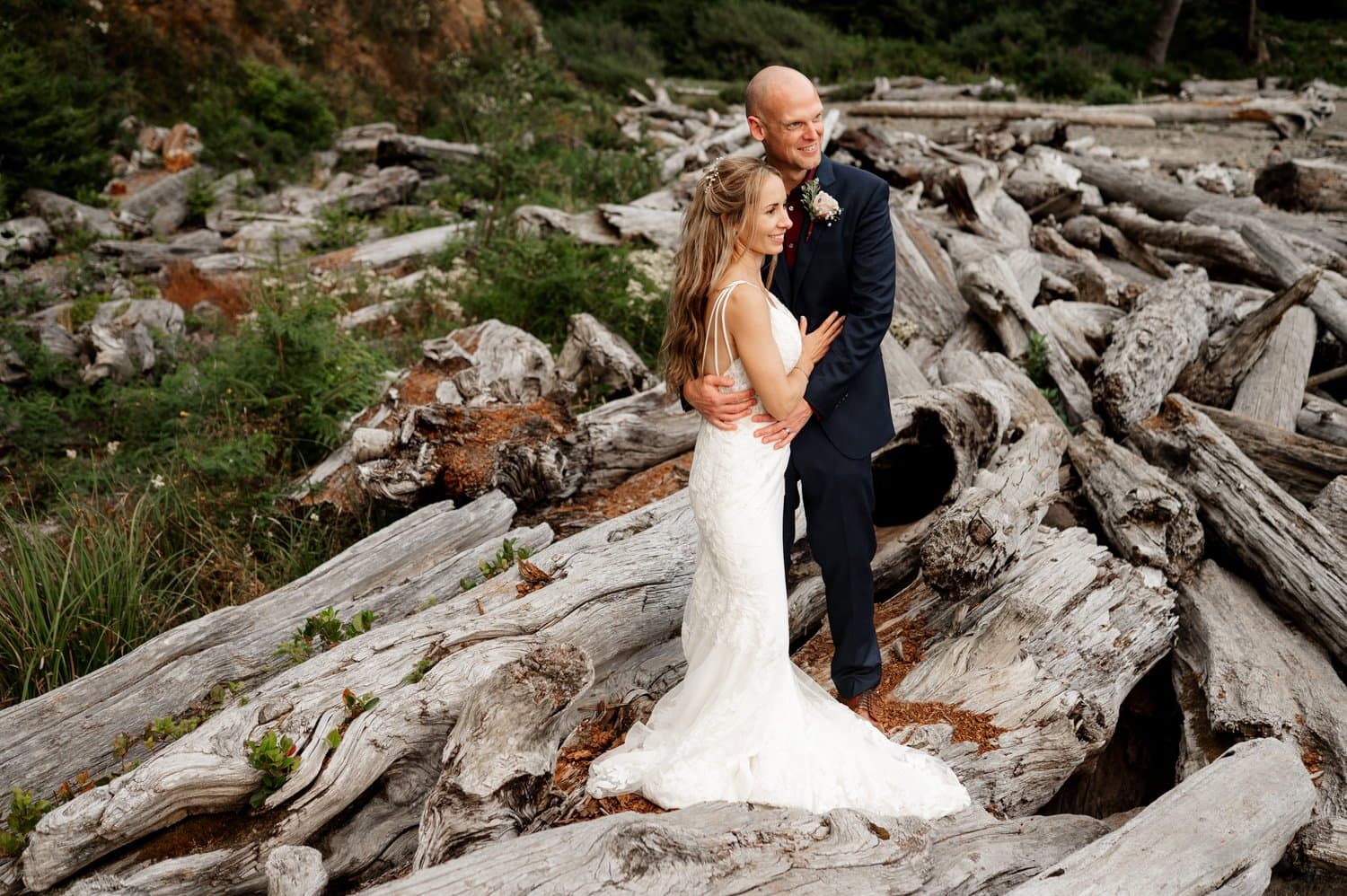 A bride in a white sleeveless gown and a groom in a dark suit embrace on large driftwood logs at Short Sands Beach. The outdoor setting boasts a natural, rustic backdrop of logs and greenery, perfect for their intimate elopement.