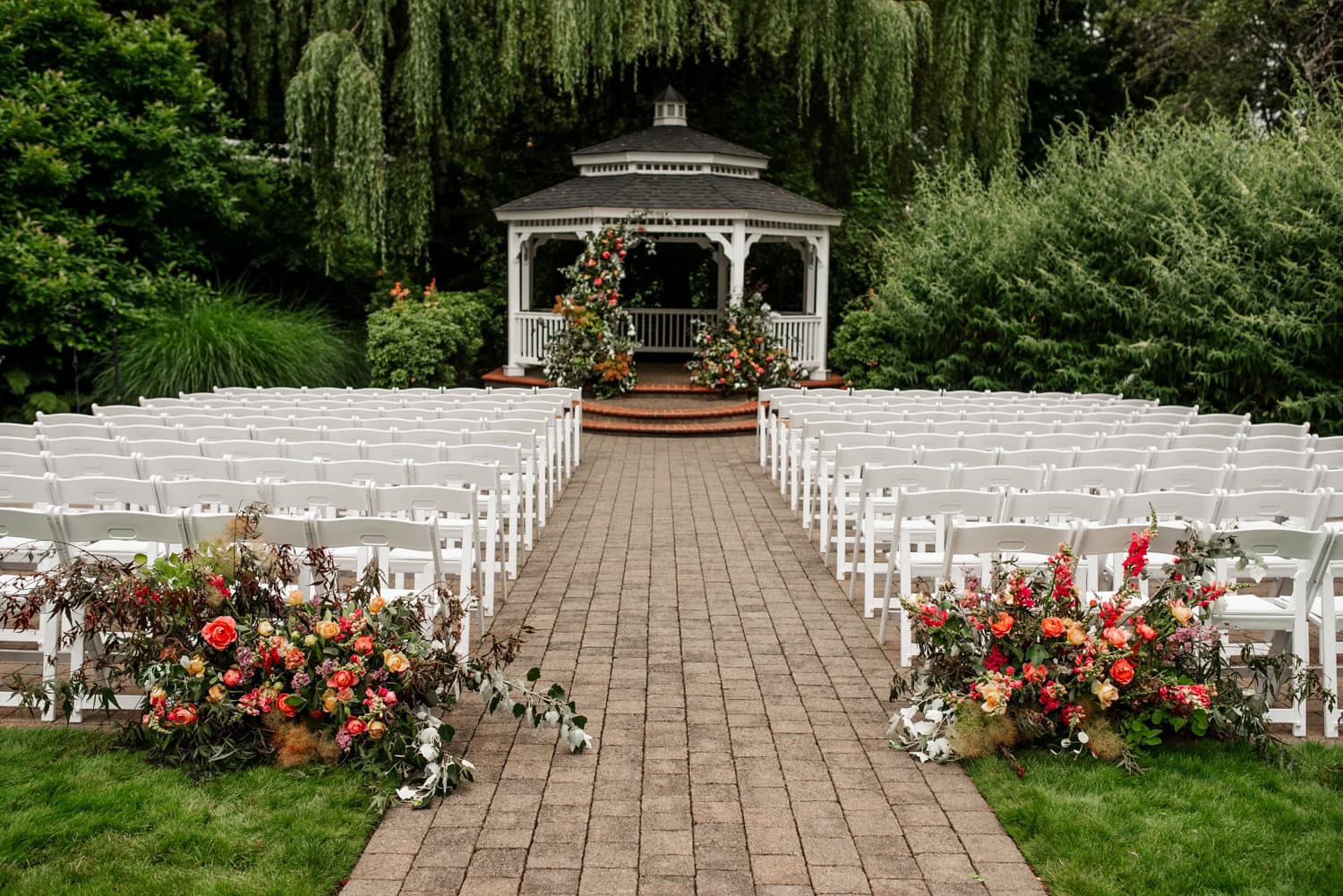 An outdoor wedding setup at Abigails Garden, Abernethy Center, features a white gazebo surrounded by greenery, a willow tree, and colorful flowers lining the aisle in Portland, Oregon.