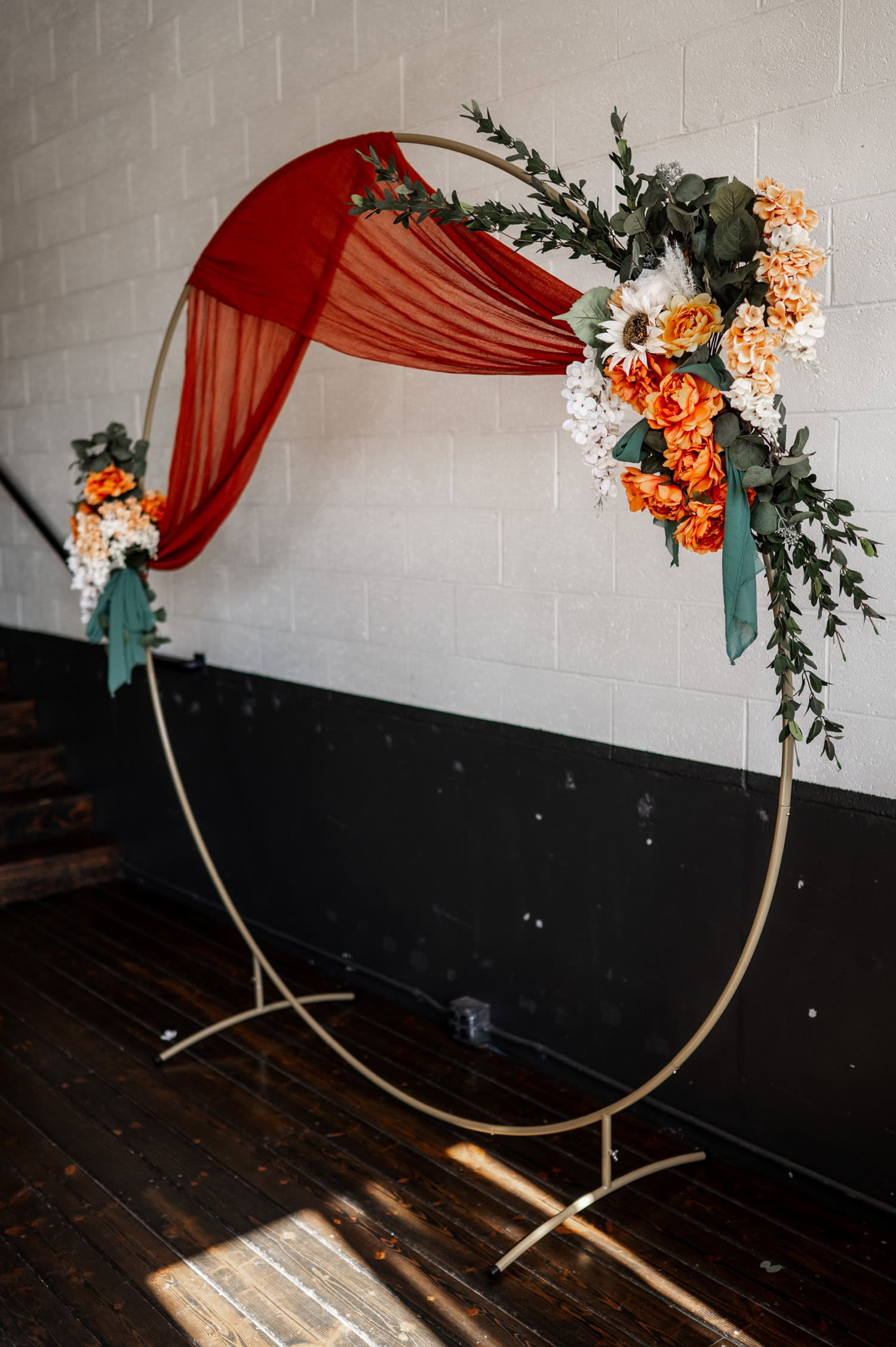A circular floral arch adorned with orange and white flowers, green foliage, and a red drape is positioned in front of a striking black and white wall at Union/Pine.