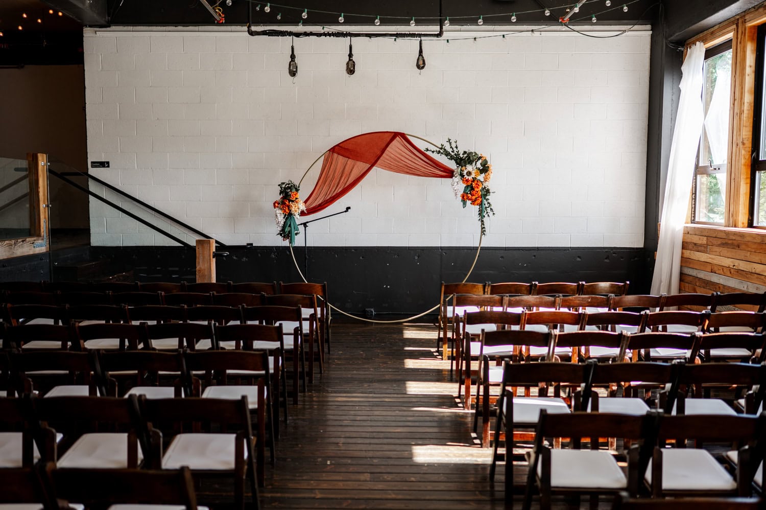 The wedding ceremony setup at Union/Pine includes wooden chairs arranged in front of a circular floral arch with string lights hanging above in a rustic room.