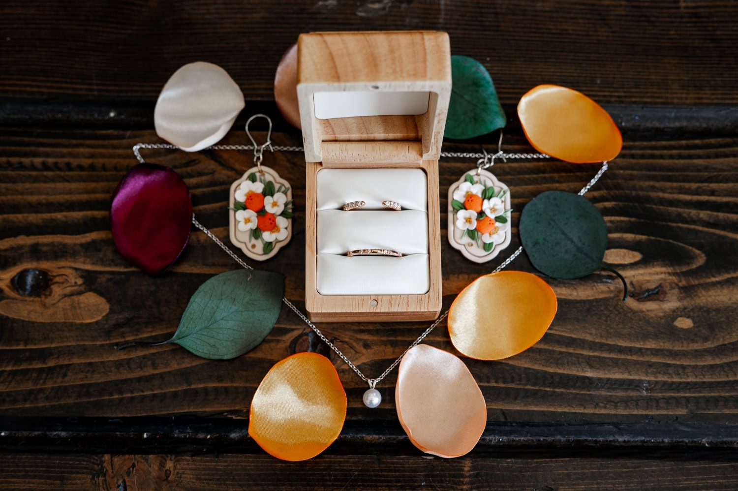A wooden ring box containing two rings is surrounded by colorful leaves, earrings, and a necklace on a rustic surface.