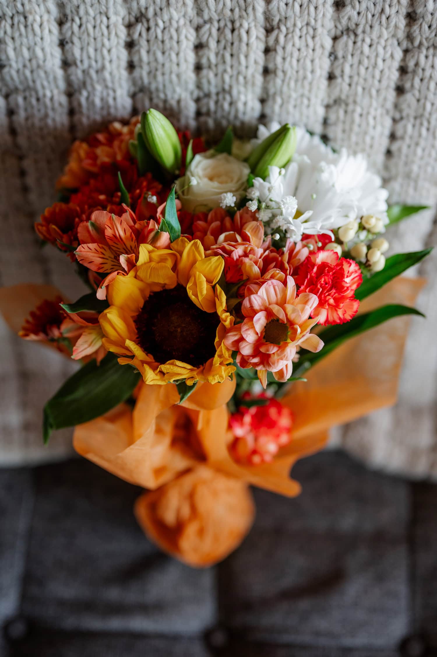 A vibrant bouquet with sunflowers, daisies, and orange flowers wrapped in orange tissue against a knitted background.