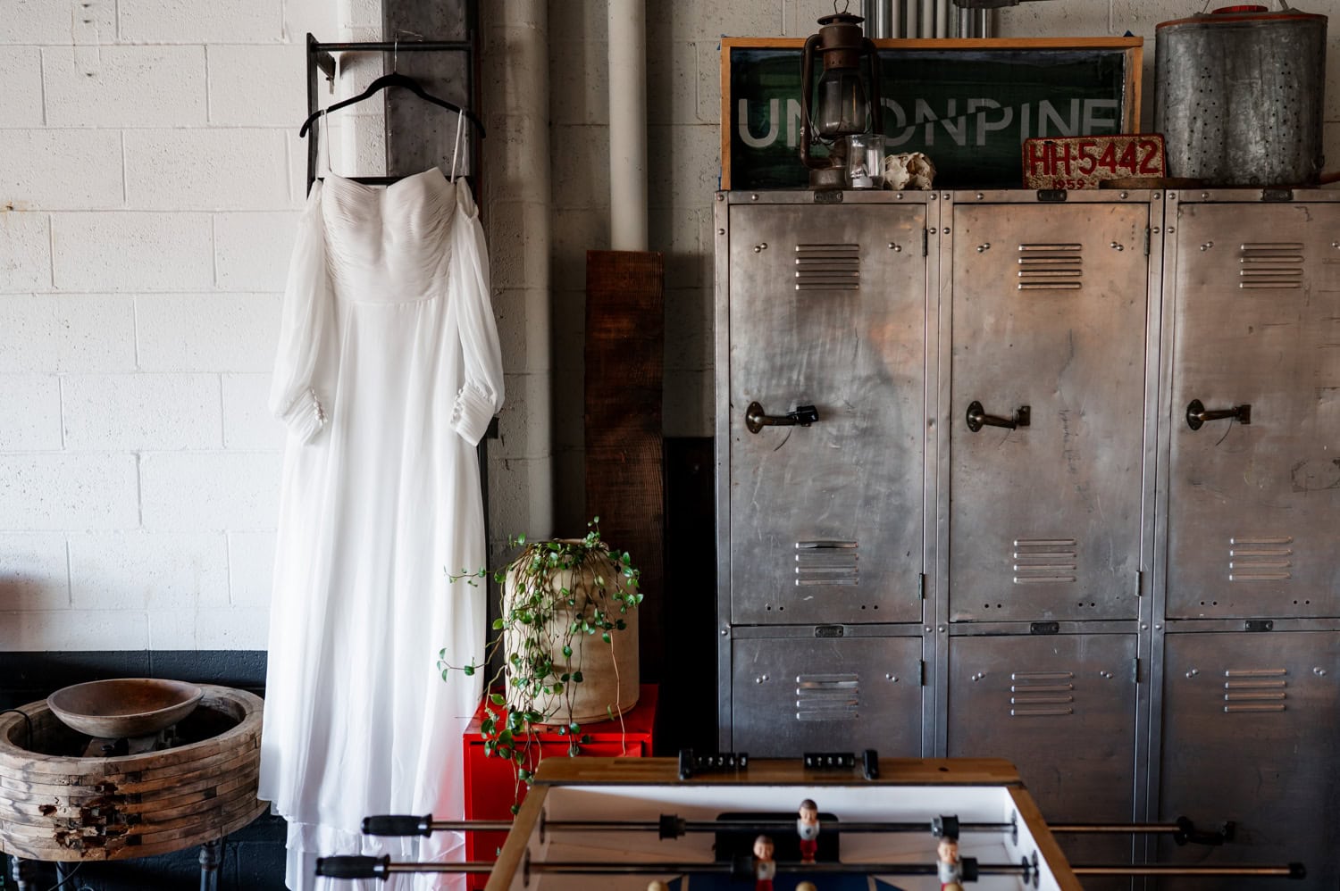 A white dress on a hanger is displayed in an industrial-looking space at Union/Pine, featuring vintage lockers, a leafy plant, and a foosball table amidst rustic decor.