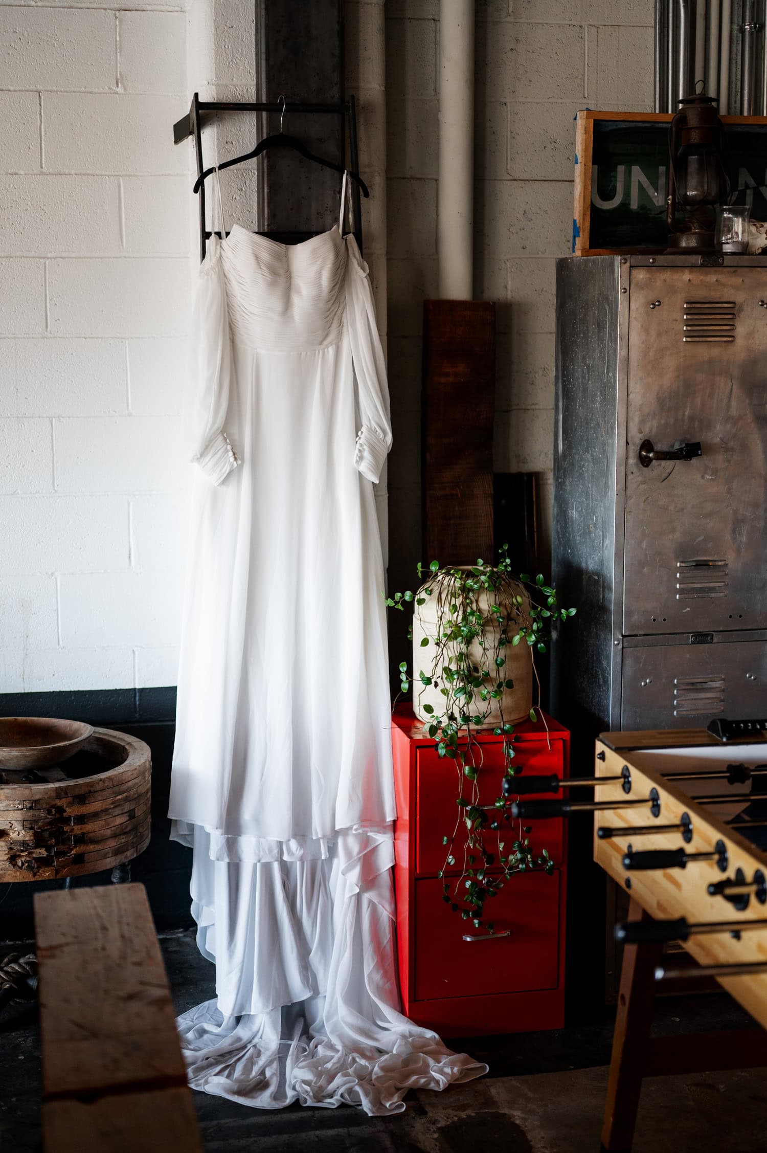 A white wedding dress is displayed in an industrial-style room at Union/Pine, next to a metal cabinet and a lush plant on a red stand.