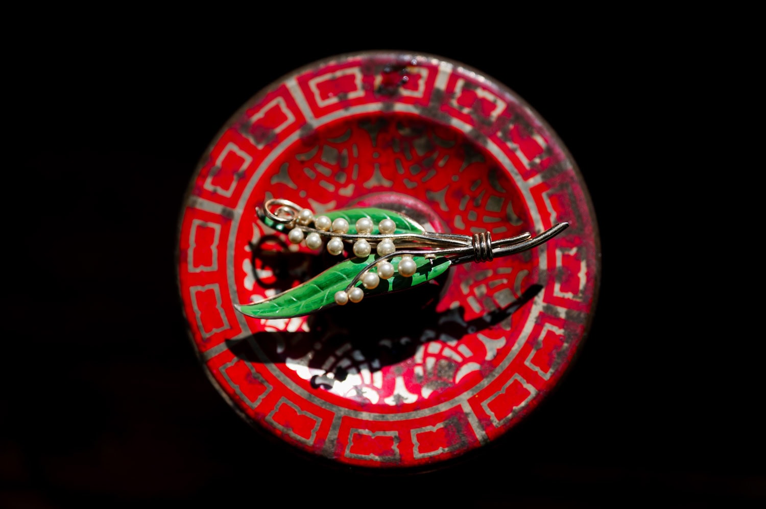 A brooch adorned with pearls and green leaves is placed on a decorative red and black circular surface.