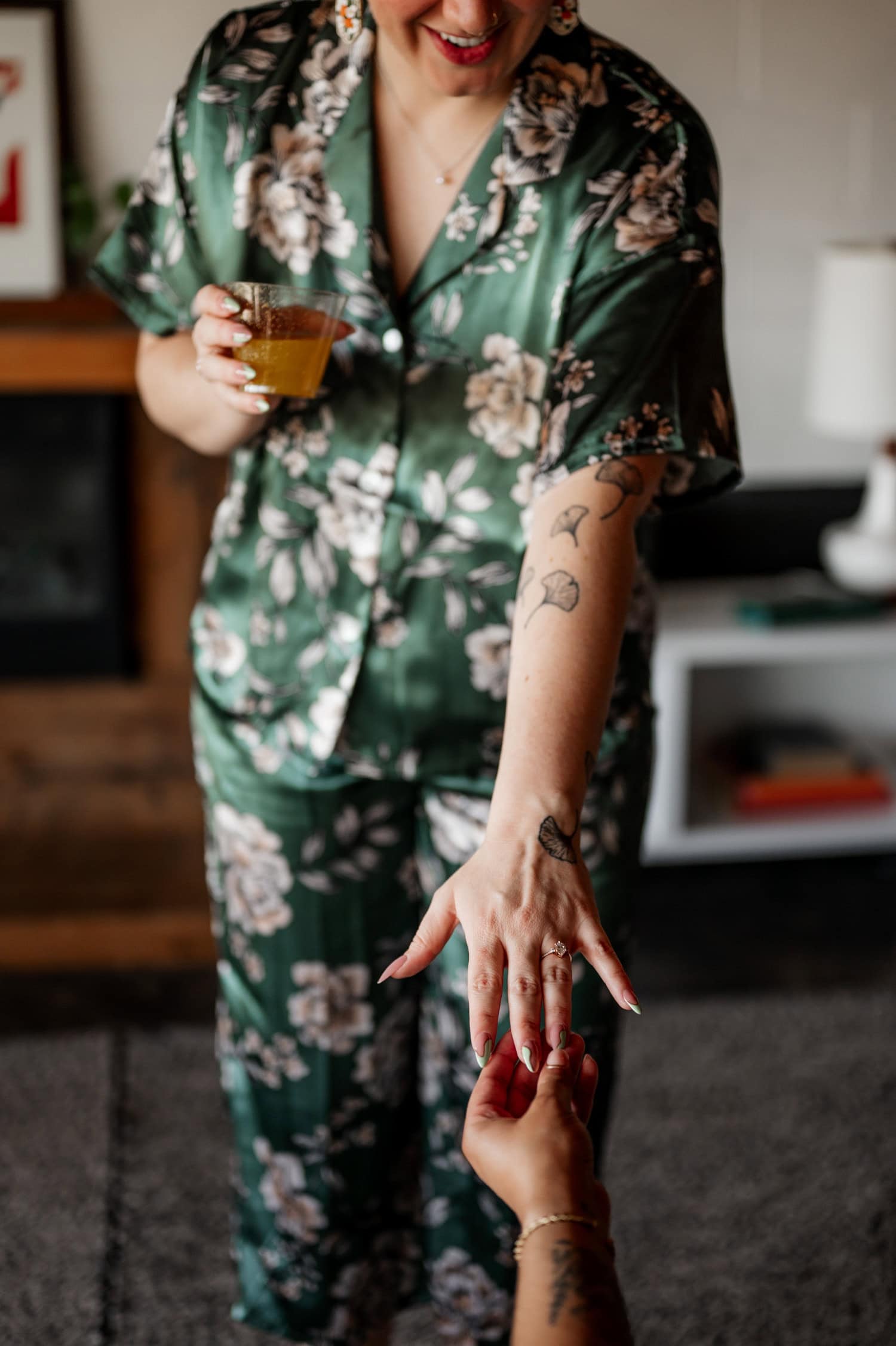 A person in floral pajamas with tattooed arms holds a drink and reaches out, standing in a stylish interior space decorated for a wedding at Union/Pine.
