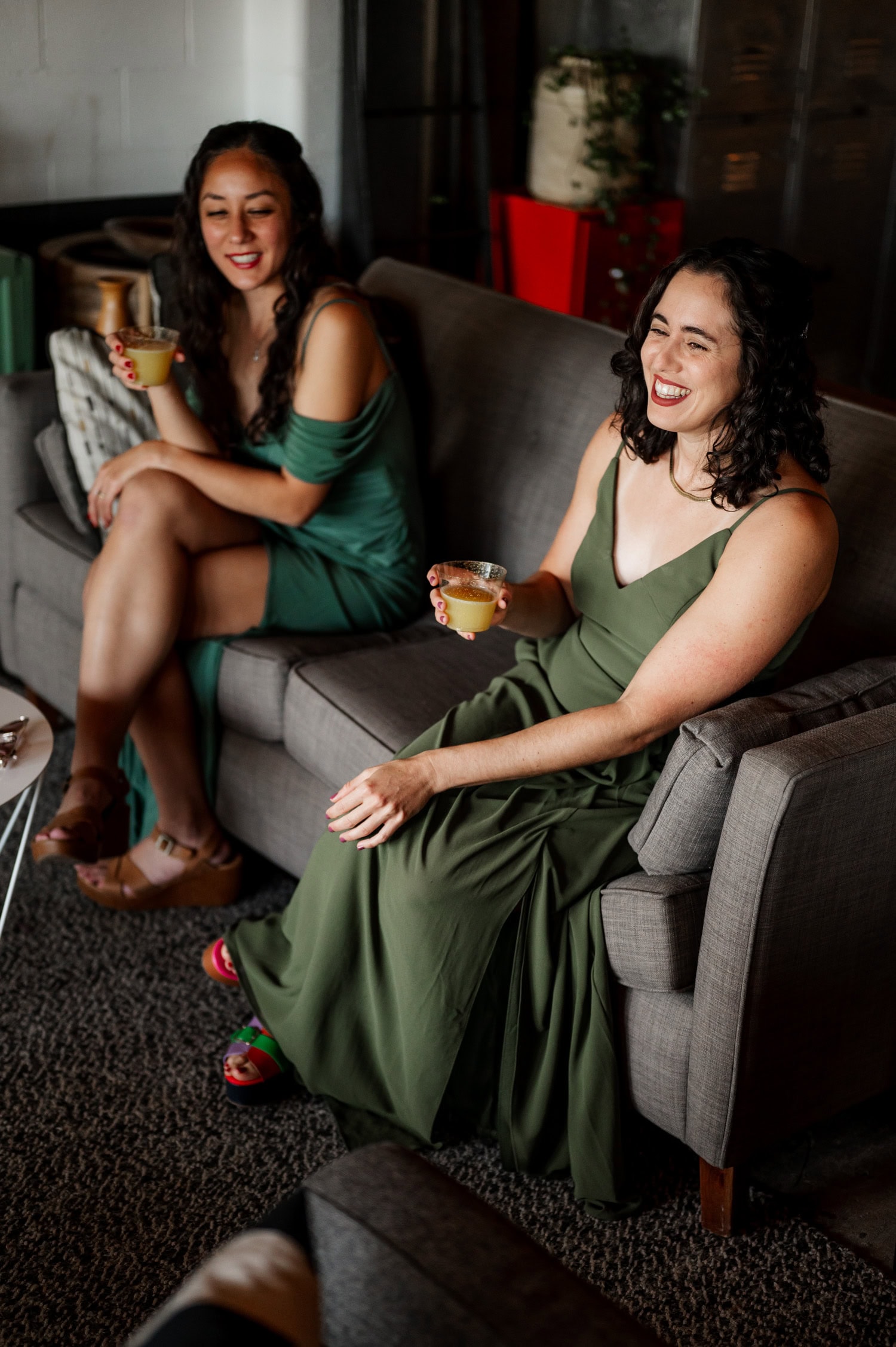 Two women in green dresses sit on a gray couch at the wedding at Union/Pine, smiling and holding drinks.