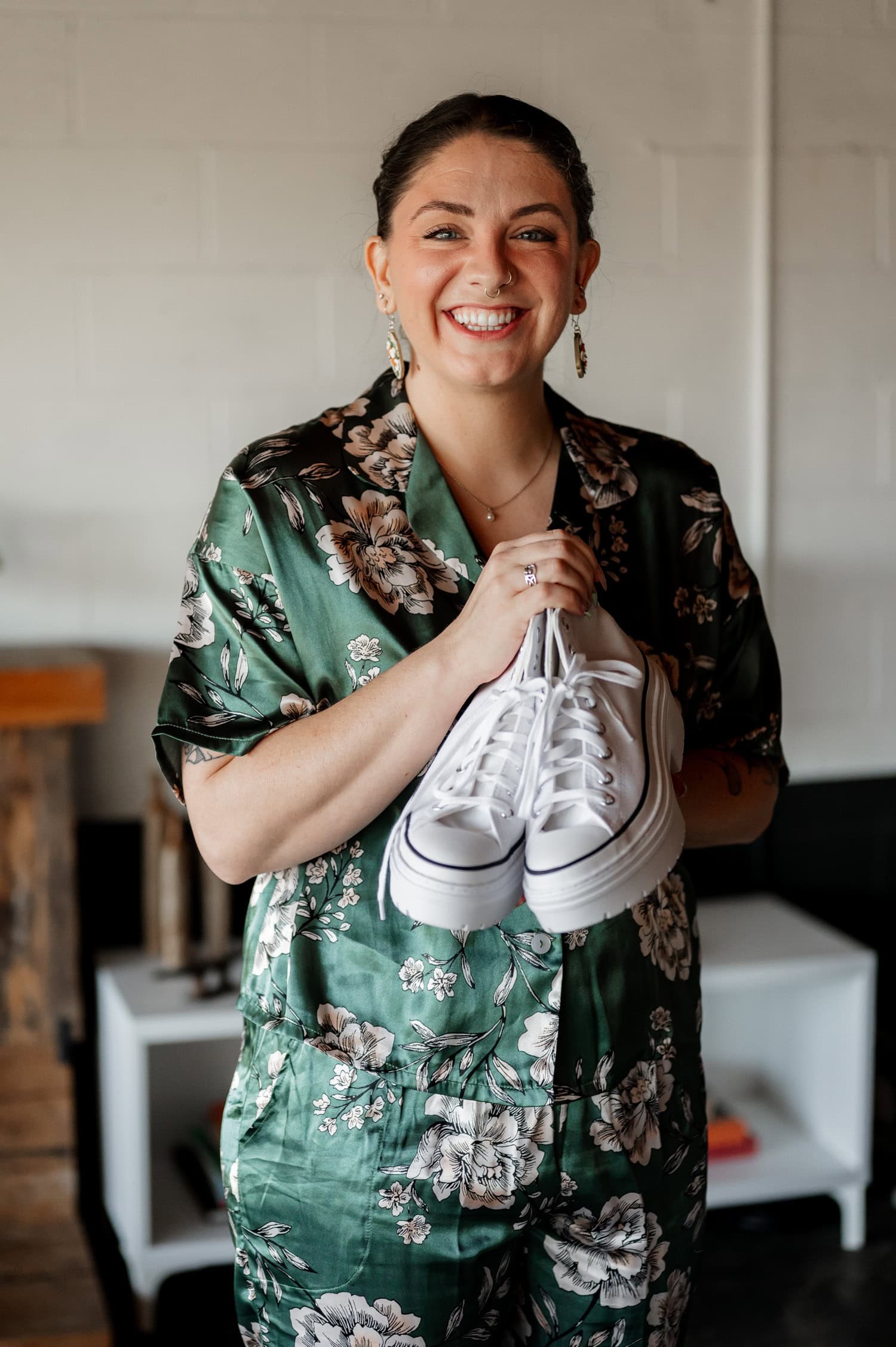 A person in a floral green outfit stands indoors, smiling and holding white sneakers. The floral pattern contrasts nicely with the background, creating a casually elegant look.