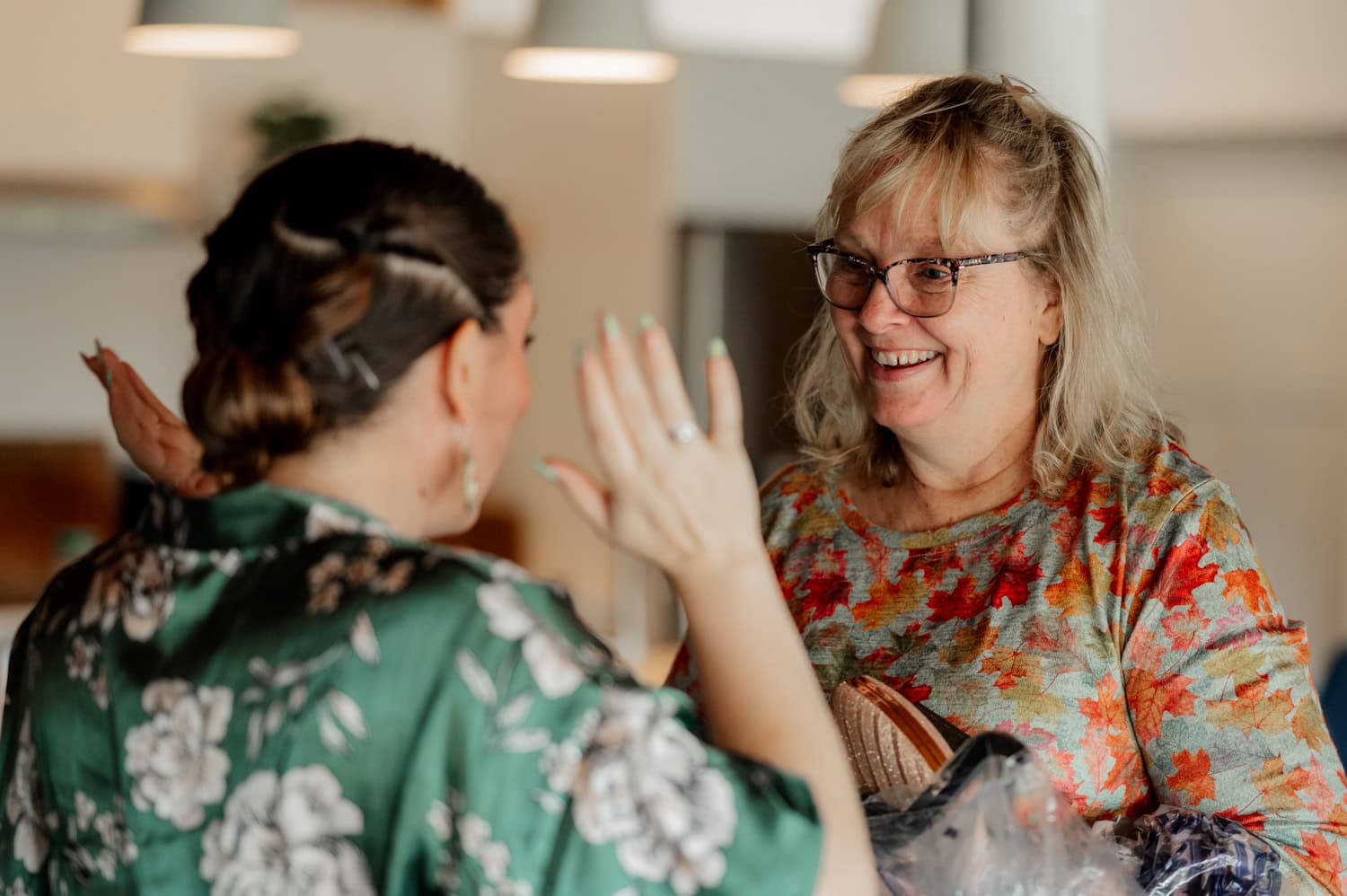 Two women are joyfully talking at a wedding at Union/Pine. One is wearing glasses and a floral shirt, while the other is dressed in green with a floral pattern.