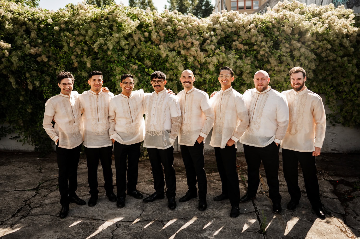 Eight men in traditional attire smiling in front of a lush green hedge at a wedding held at Union/Pine.