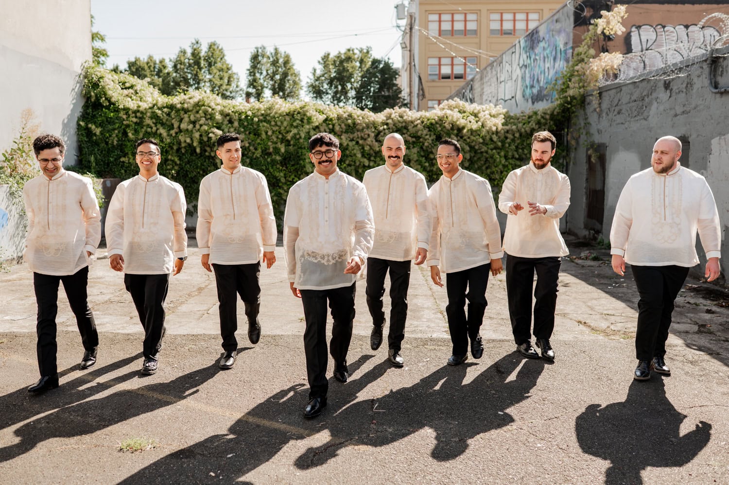 Eight men wearing traditional sheer barong shirts and dark pants smile together, enjoying a sunny day at Union/Pine, possibly for a wedding celebration.