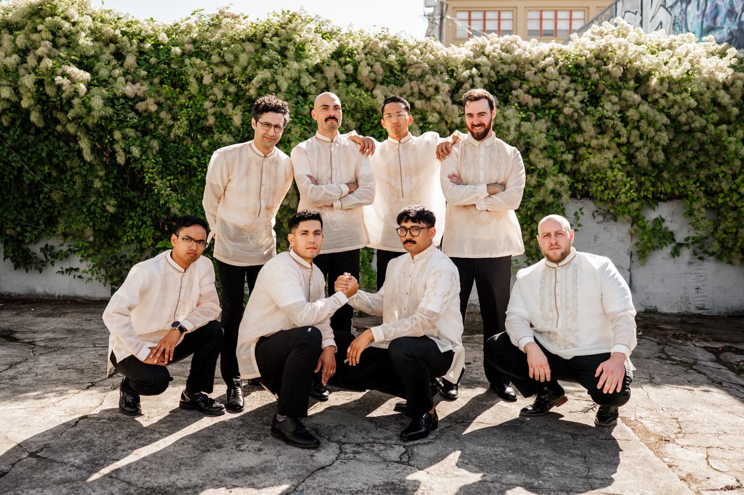A group of men in traditional attire posing outdoors, with lush greenery in the background.