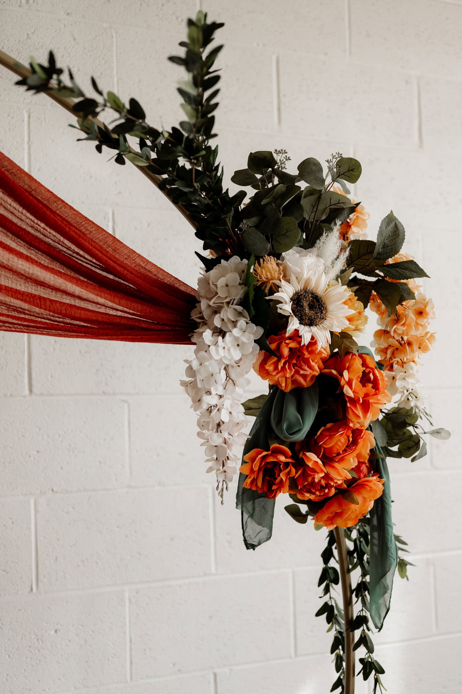 A stunning floral arrangement with orange and white flowers intertwined with lush greenery drapes against a textured white wall on red fabric at Union/Pine.