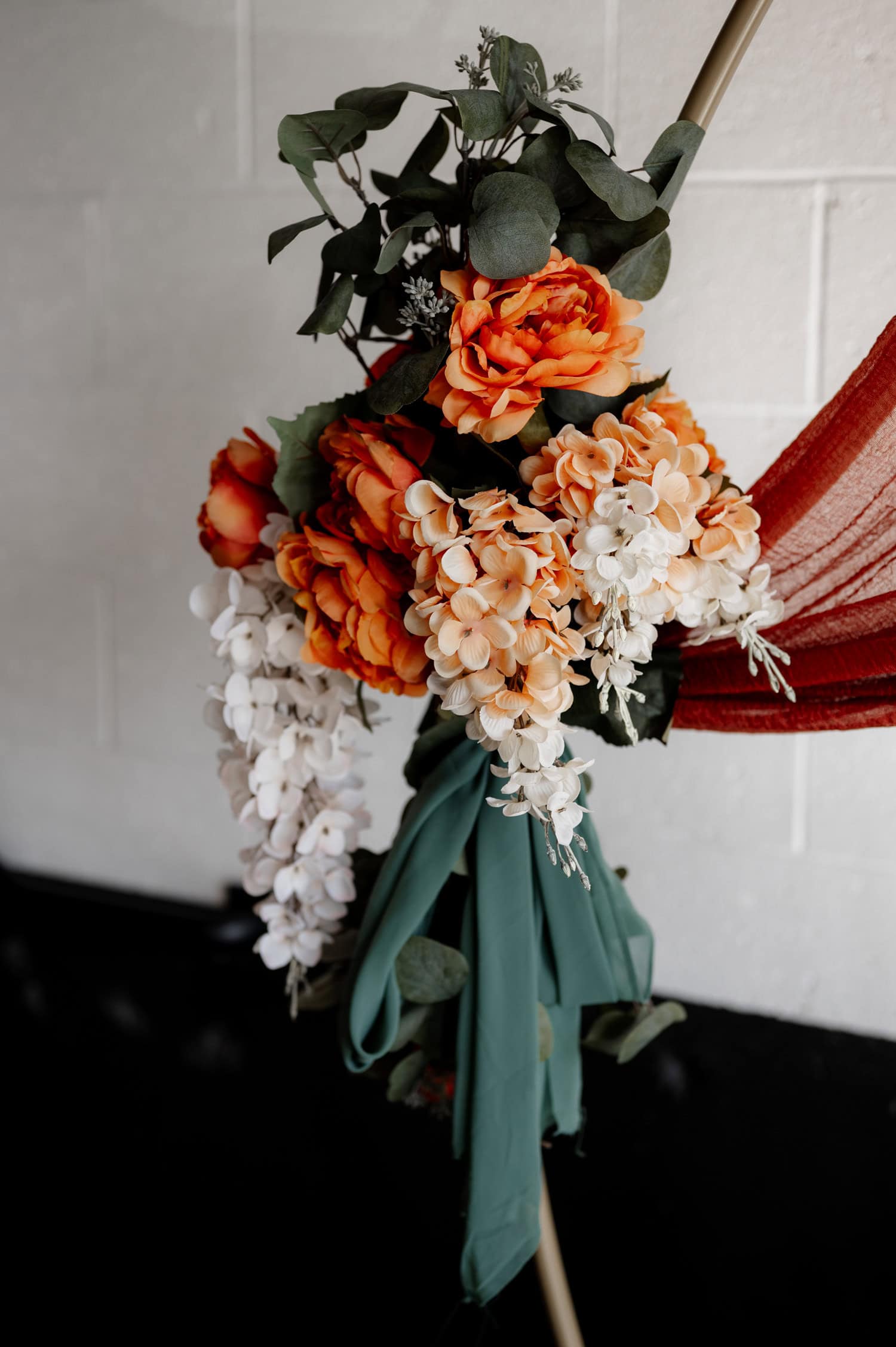 A wedding floral arrangement with orange roses, white flowers, and green leaves is adorned with draped red and green fabric.