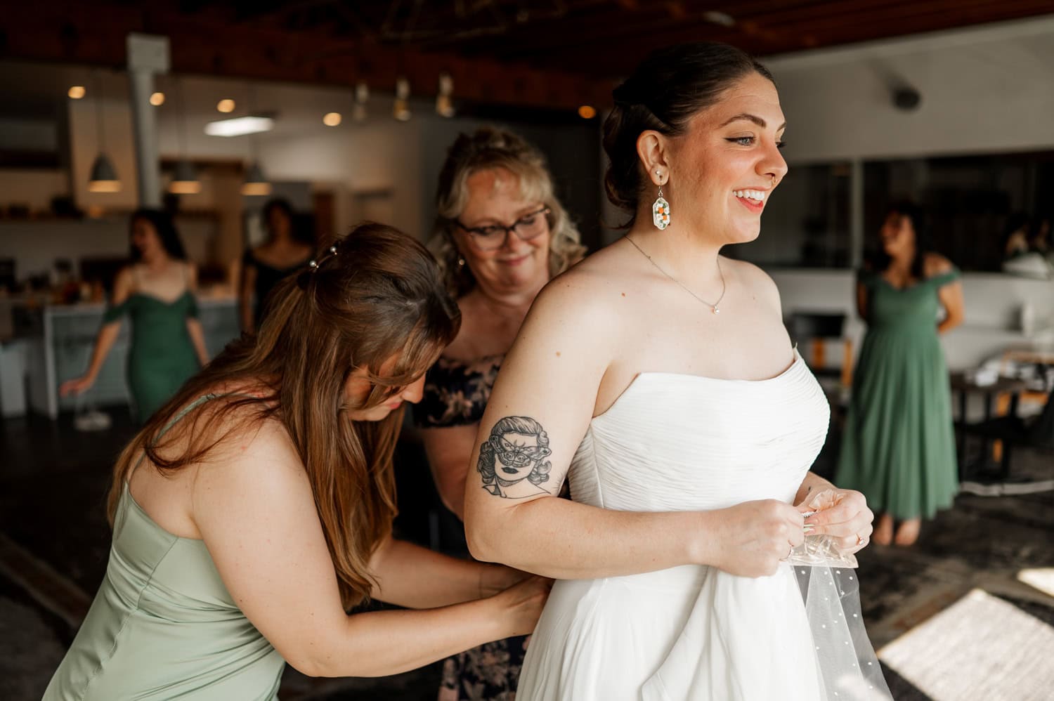 In a warmly lit room, the bride is being assisted by two women as she puts on her elegant white dress, preparing for her wedding at Union/Pine.