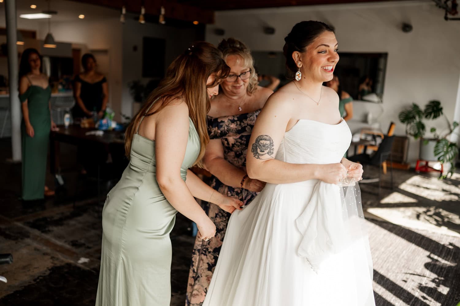 In a warmly lit room at Union/Pine, the bride smiles brightly as two women help adjust her dress. Bridesmaids are gathered in the background, chatting and looking on.