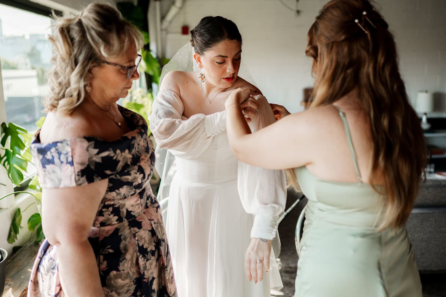 In an indoor setting with plants, a bride in a white dress adjusts her sleeve with assistance from two women in floral and green dresses. The scene captures a moment from a wedding at Union/Pine.