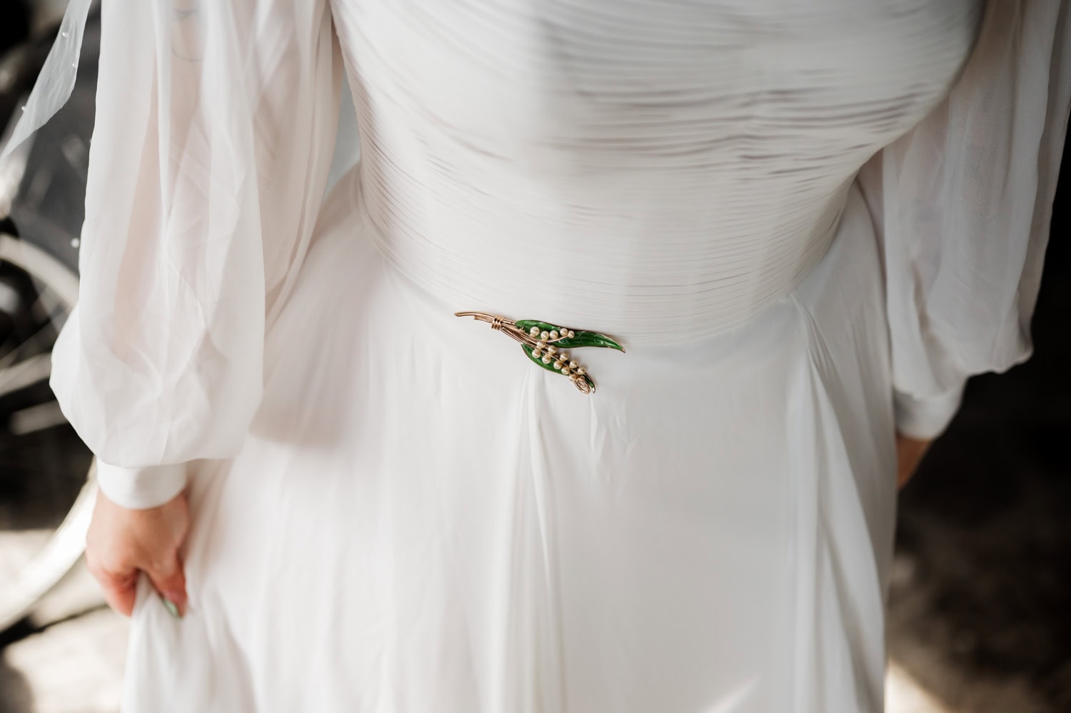 A person in a white dress with a green and gold flower brooch on the waist, standing with hands by their sides, during a wedding at Union/Pine.